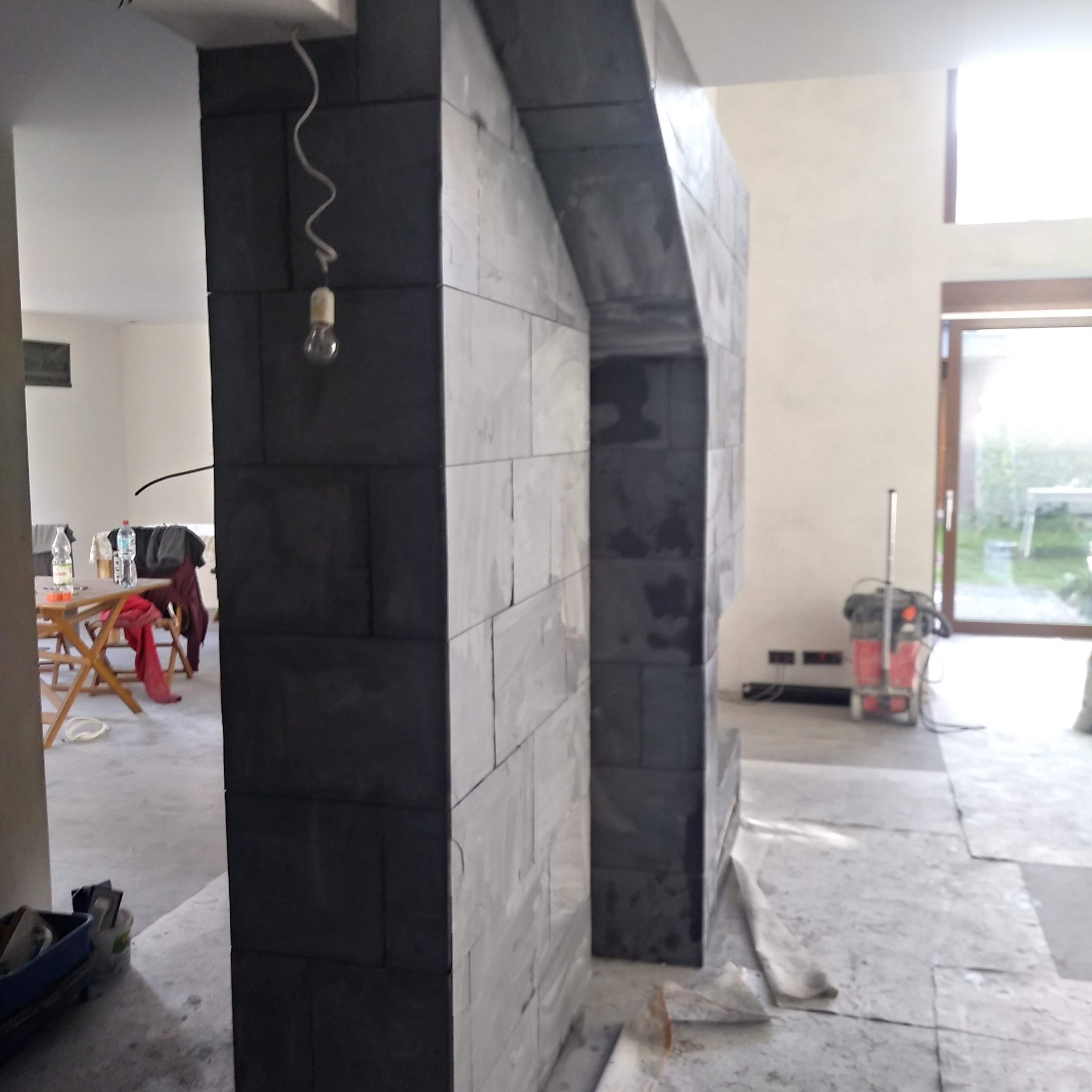 Wnętrze w trakcie wykańczania: fragment ściany z szarej, prostokątnej okładziny kamiennej tworzącej łuk, widoczny fragment podłogi z płyt, narzędzia w pojemniku, lampa wisząca na kablu i odkurzacz...