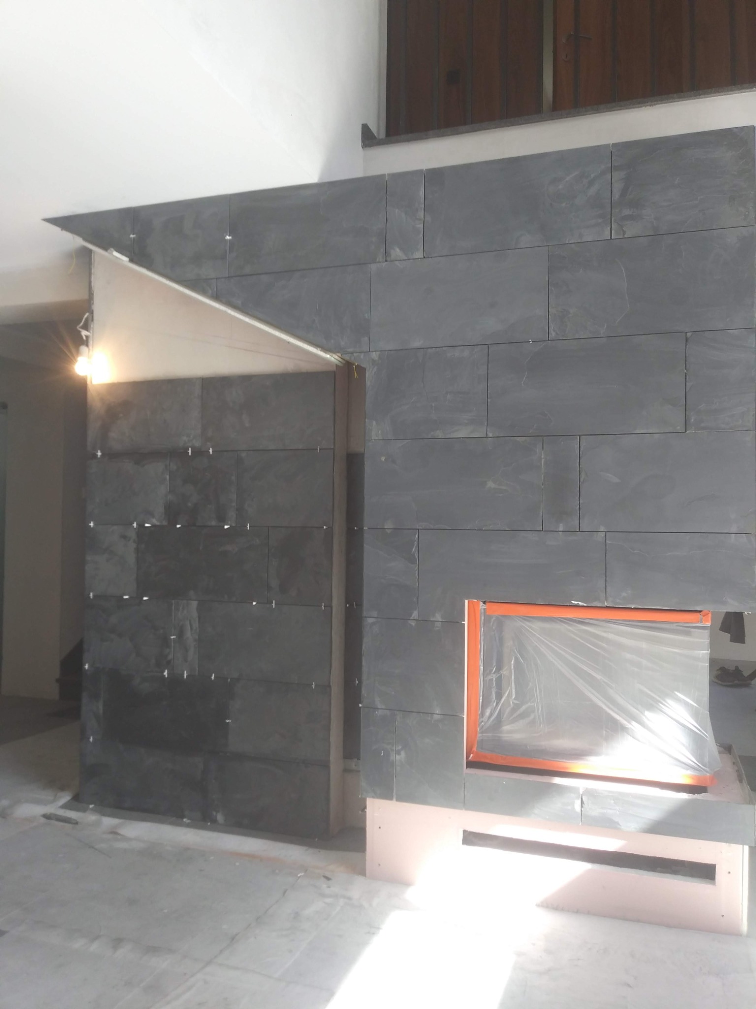 Montaż ciemnoszarych płytek na ścianie kominka, widoczne krzyżyki dystansowe, zabezpieczone folią otwór kominkowy i fragment sufitu.