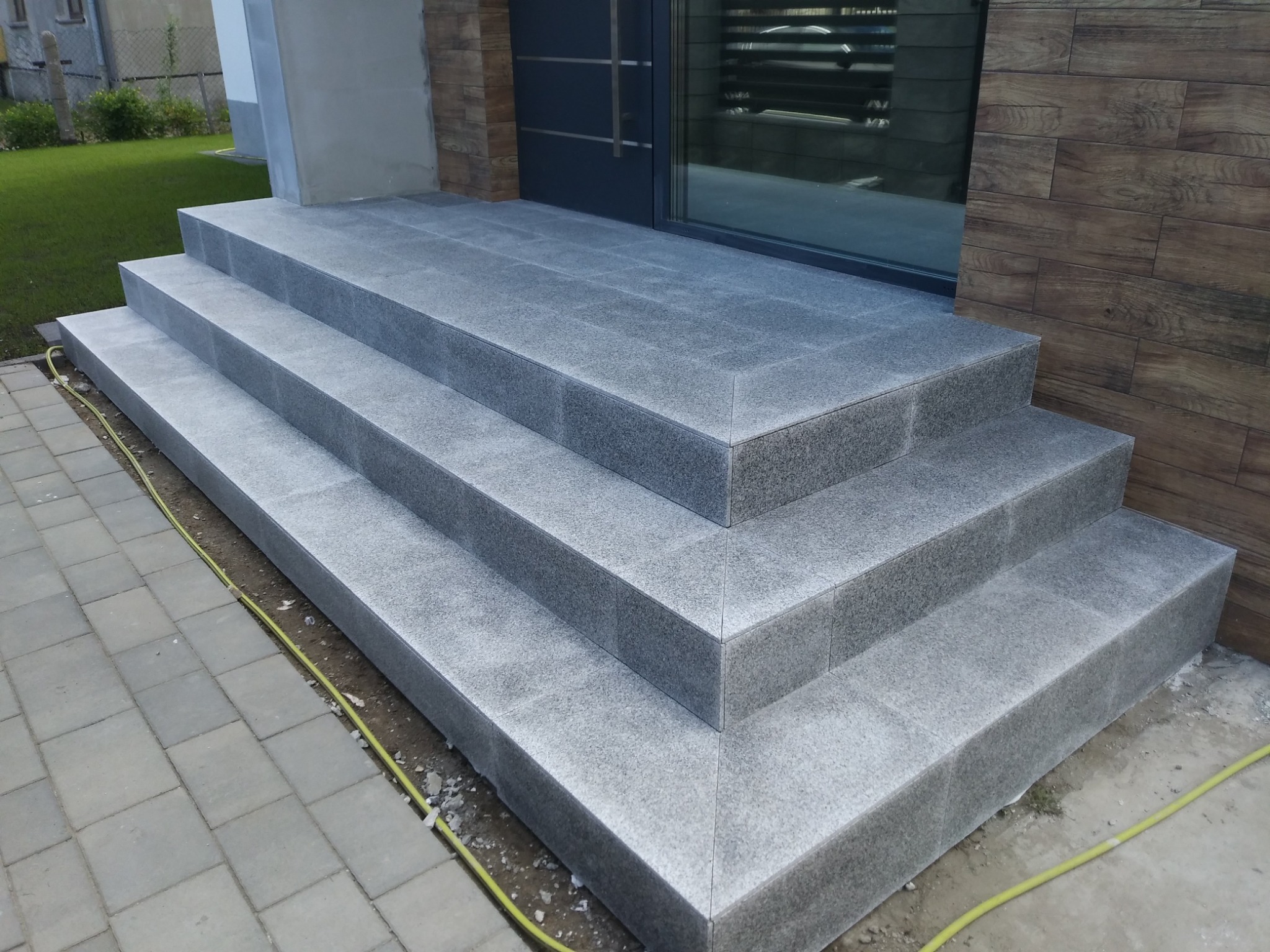 Zewnętrzne schody wejściowe wyłożone szarymi płytami granitowymi, widoczny fragment elewacji z drewnopodobnych paneli i trawnik w tle.