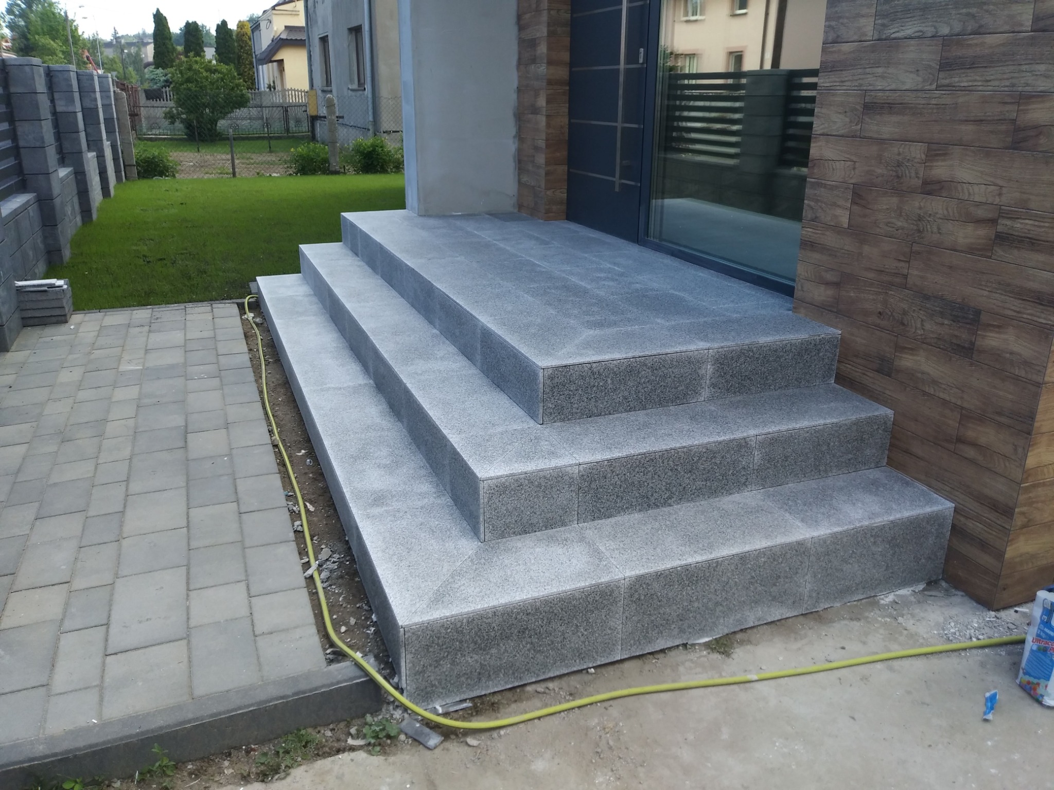 Granitowe schody wejściowe do domu z szarą stolarką okienną i drzwiową, wykończone drewnopodobną okładziną, widoczny fragment trawnika i kostki brukowej.