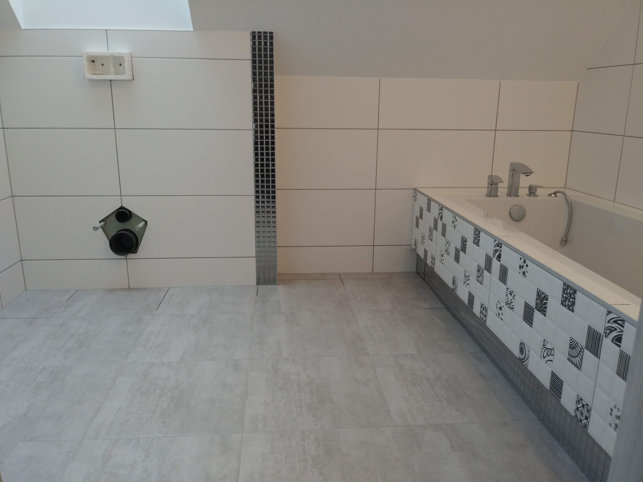 Wnętrze łazienki w trakcie wykańczania, widoczne białe płytki ścienne z czarnym paskiem dekoracyjnym, szare płytki podłogowe, wanna z obudową dekorowaną wzorzystymi płytkami, oraz niezamaskowane...