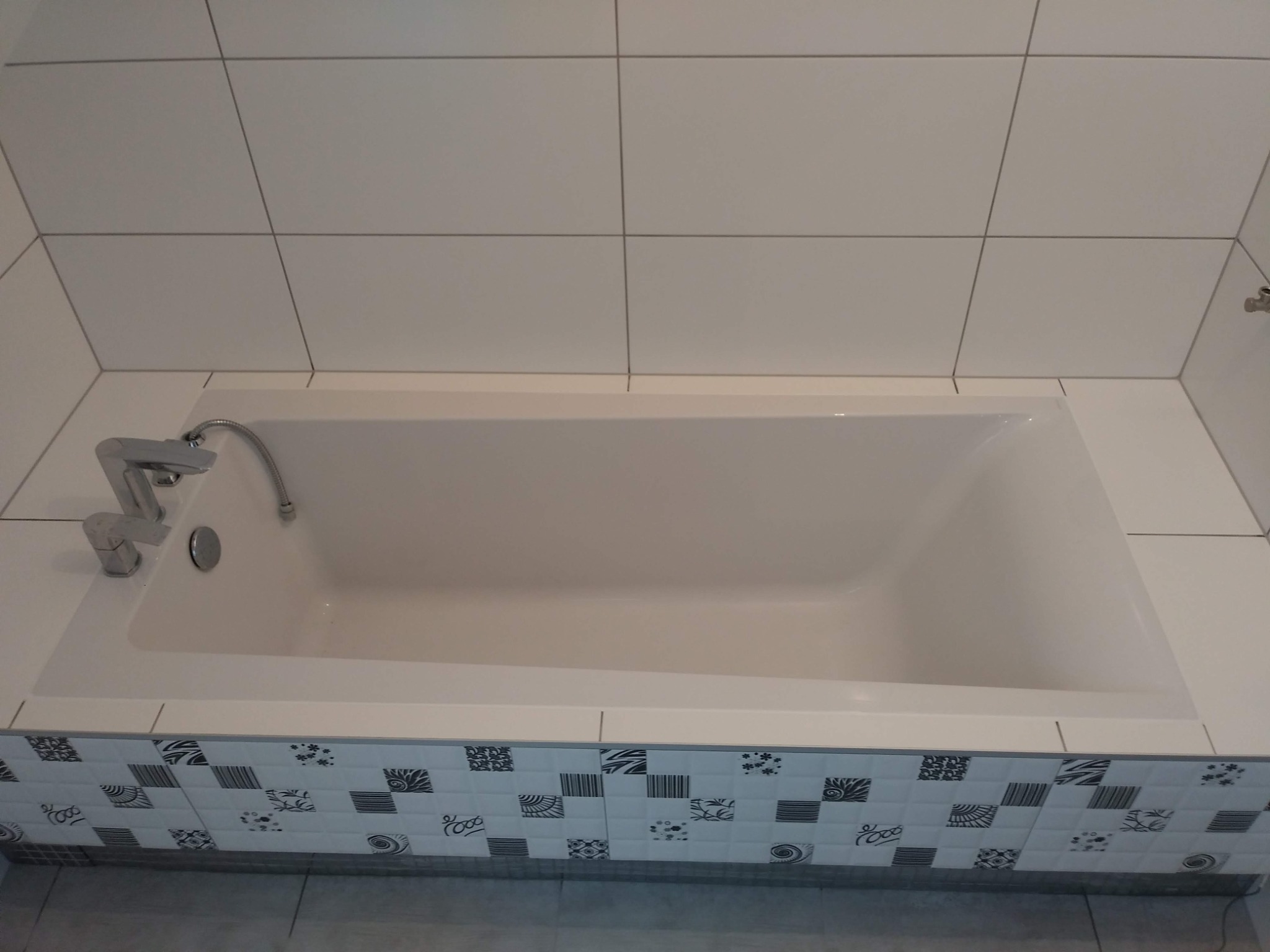 Biała wanna obudowana płytkami z czarno-białym wzorem, armatura chromowana, ściany wyłożone białymi, kwadratowymi płytkami.