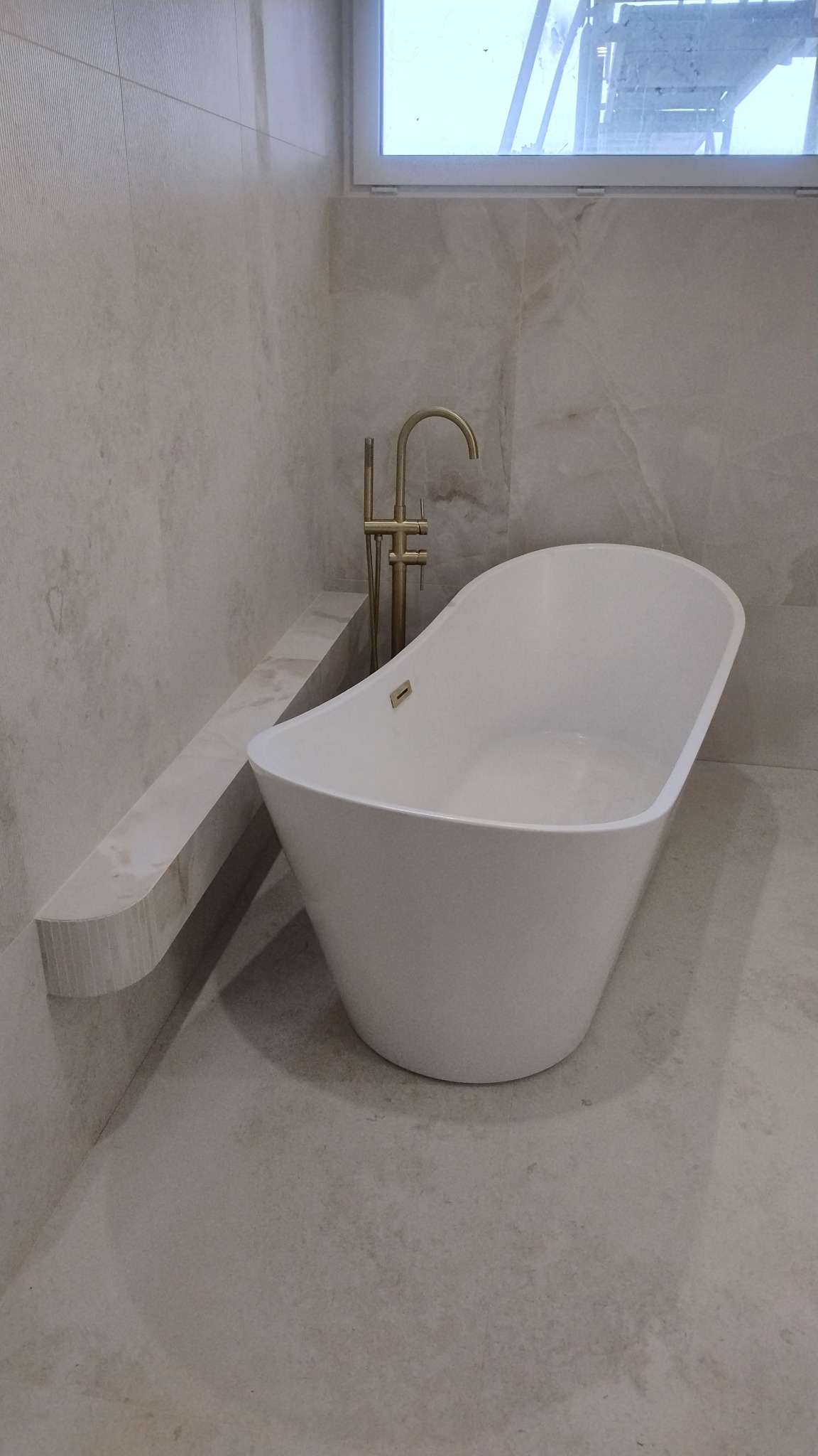 Elegancka, wolnostojąca wanna w odcieniach bieli i beżu, z baterią w kolorze starego złota, umieszczona w wykończonej łazience z marmurowymi płytkami.