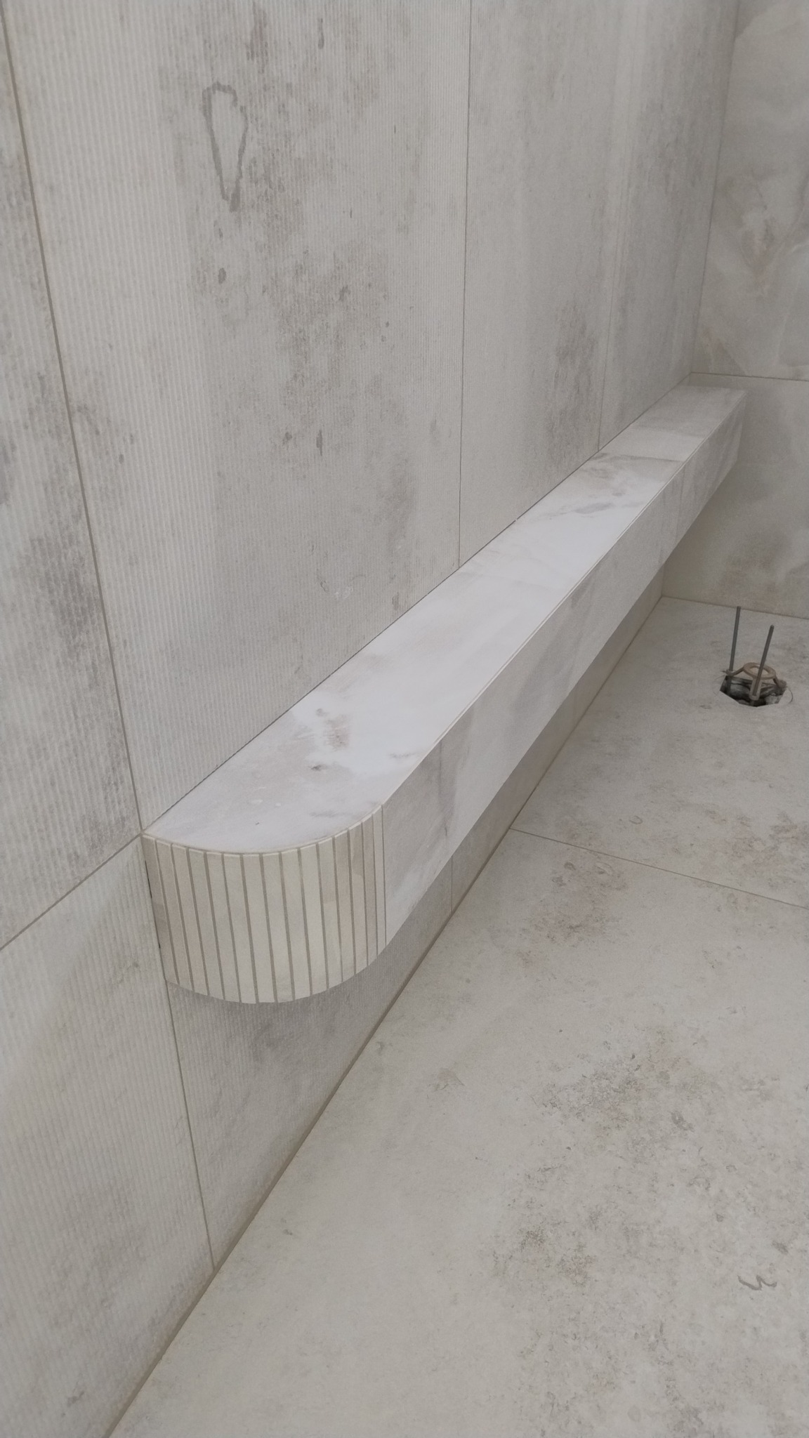 Wykończona półka z płytek w kabinie prysznicowej, z zaokrąglonym frontem i subtelnym wzorem. Widoczne płytki na ścianach i podłodze w odcieniach szarości.