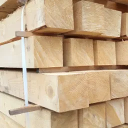 Stos świeżo przetartego drewna konstrukcyjnego, jasne, kwadratowe belki ułożone warstwami, zabezpieczone paskami i przekładkami, widoczne słoje drewna.