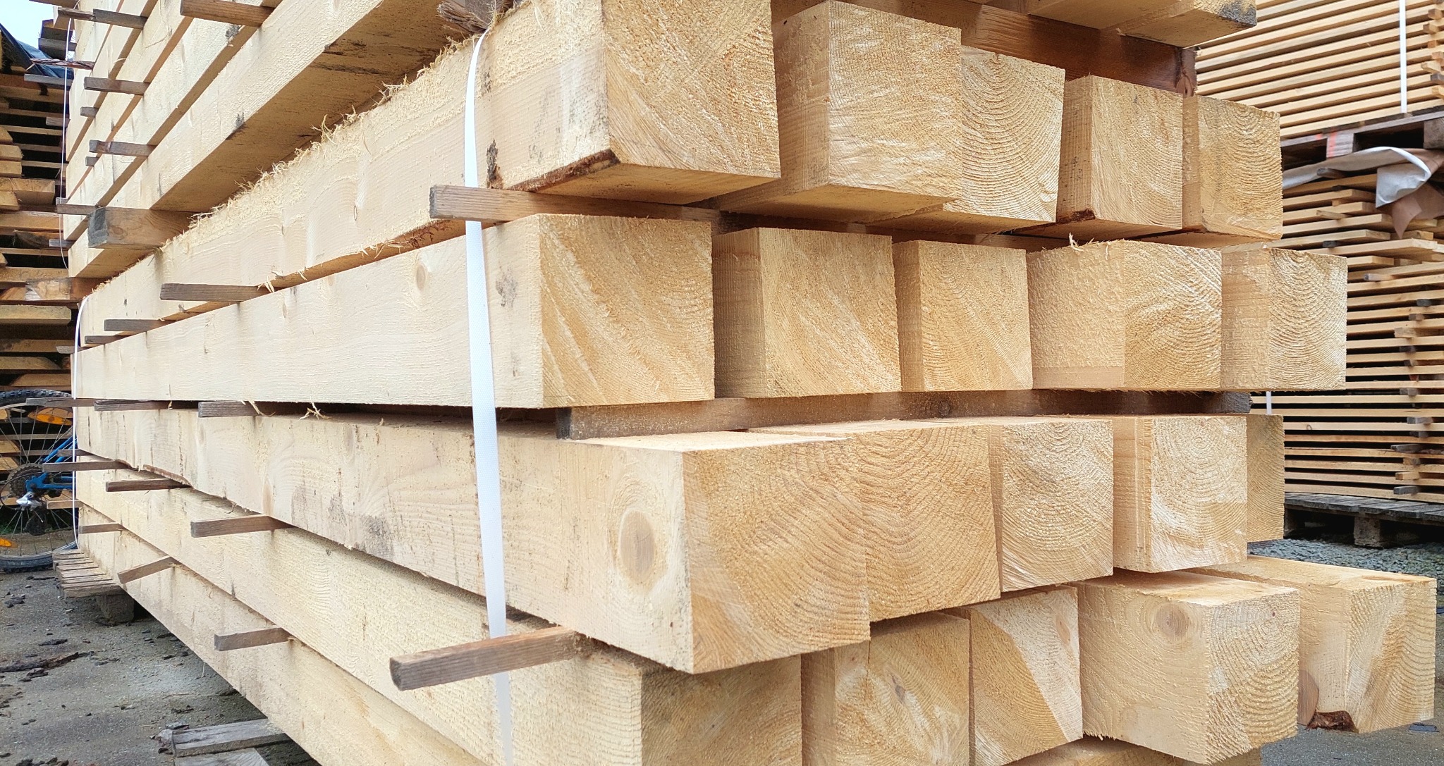 Stos świeżo przetartego drewna konstrukcyjnego, jasne, kwadratowe belki ułożone warstwami, zabezpieczone paskami i przekładkami, widoczne słoje drewna.