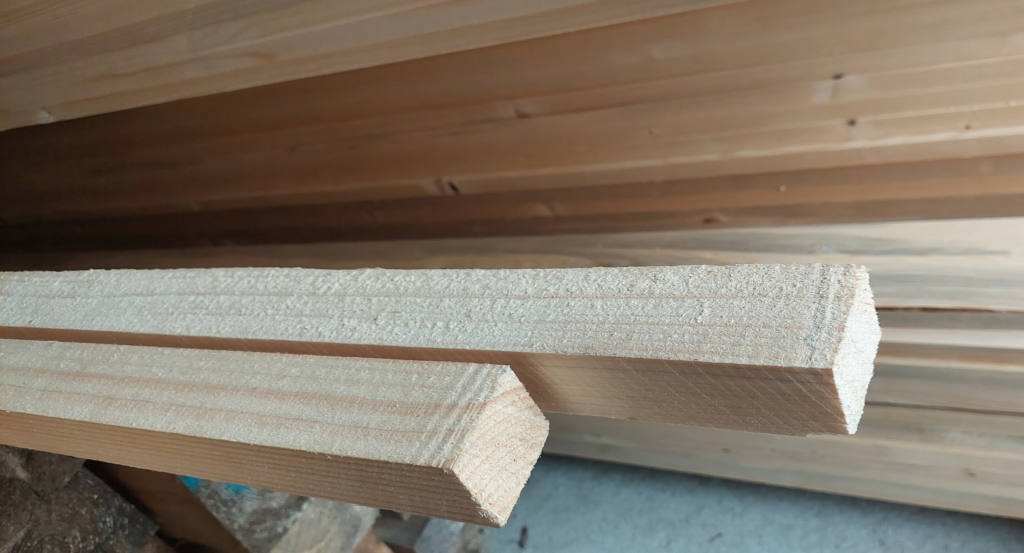 Bliski kadr dwóch kantówek z jasnego drewna, lekko zakurzonych, ułożonych jedna nad drugą na tle stosu desek.