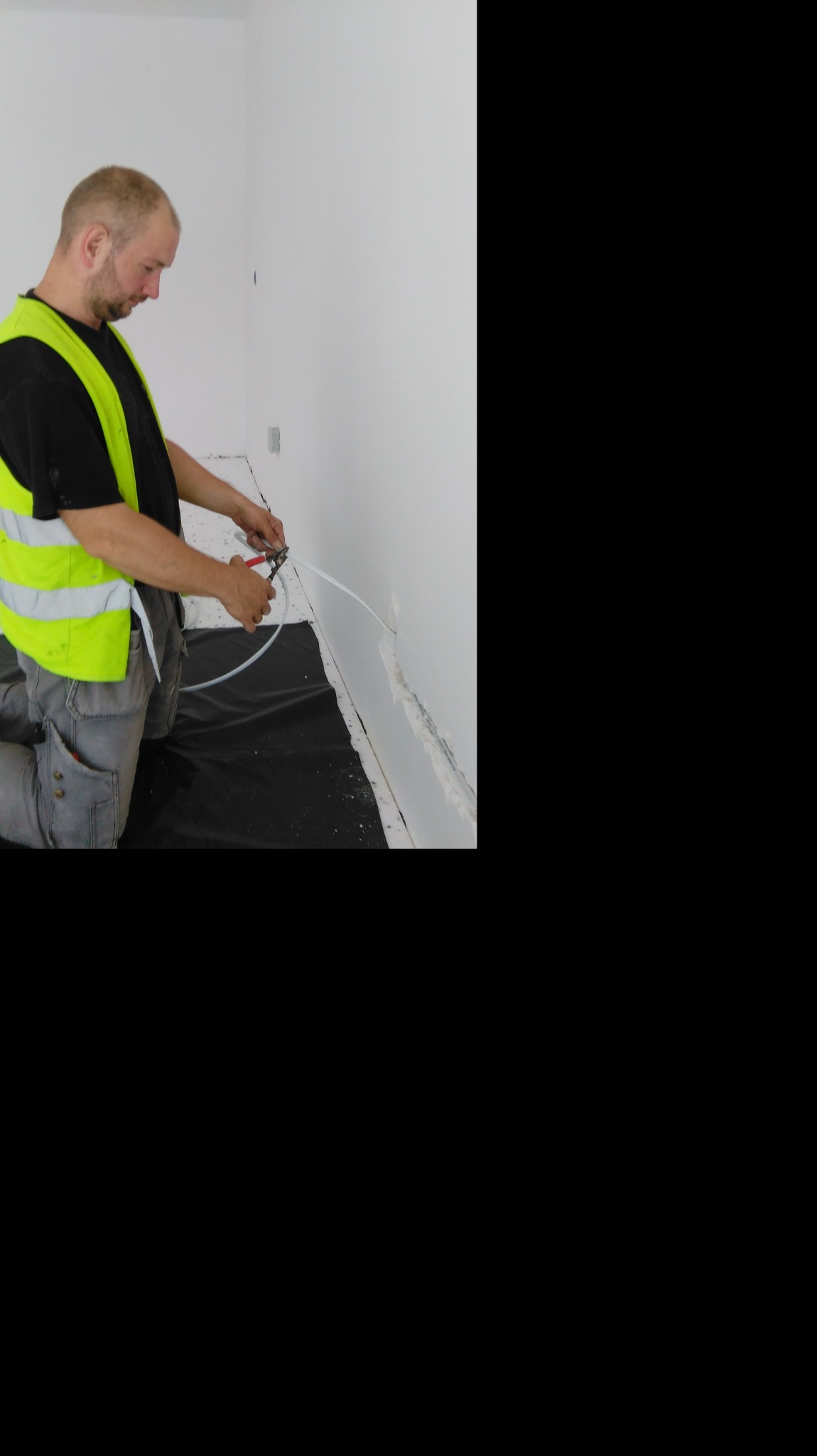Elektryk w kamizelce odblaskowej przycina przewód elektryczny przy ścianie podczas prac remontowych, zabezpieczona podłoga.