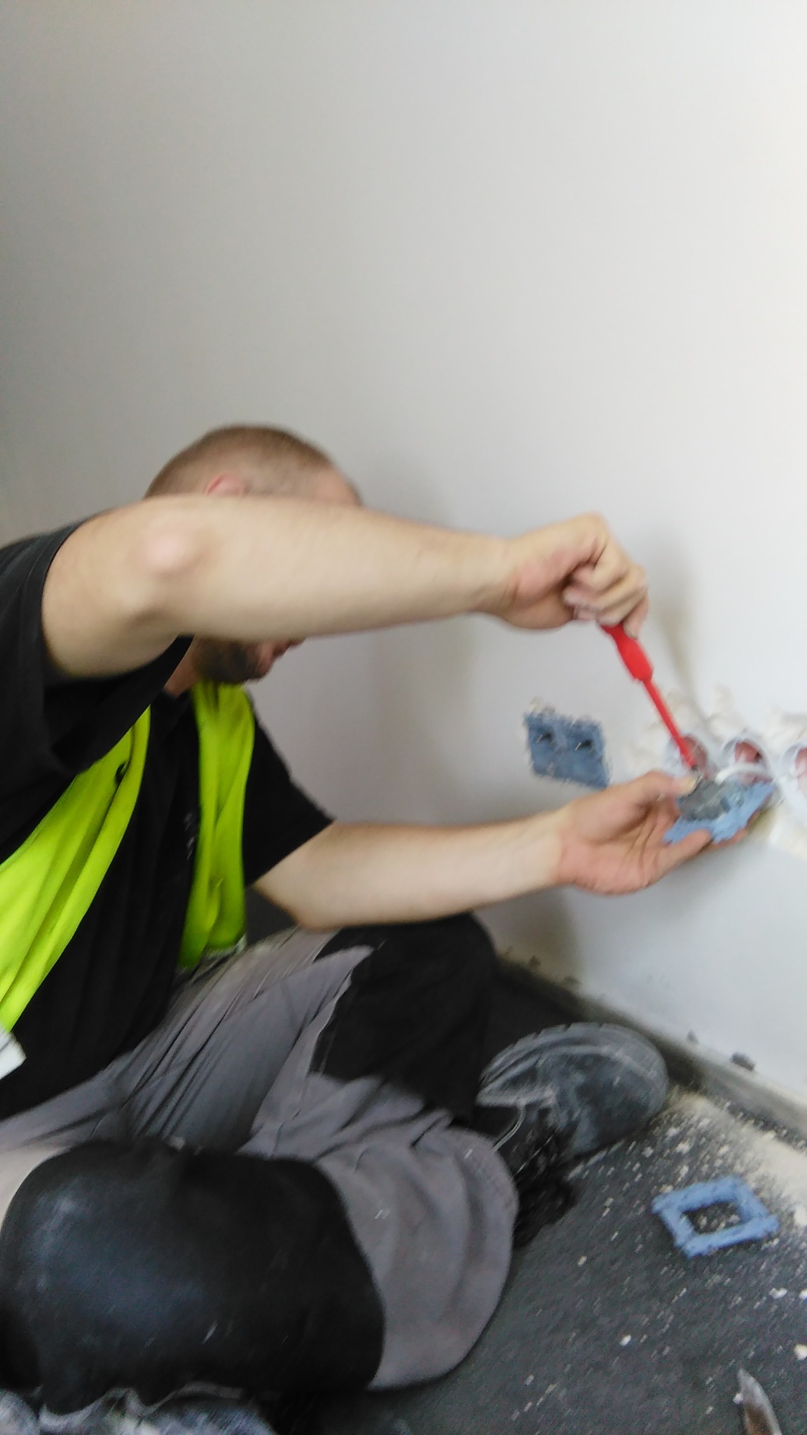 Elektryk w kamizelce odblaskowej, czyszczący gniazdo elektryczne z resztek zaprawy czerwoną szpachelką, widoczne puszki instalacyjne w ścianie i drobne zabrudzenia na podłodze.
