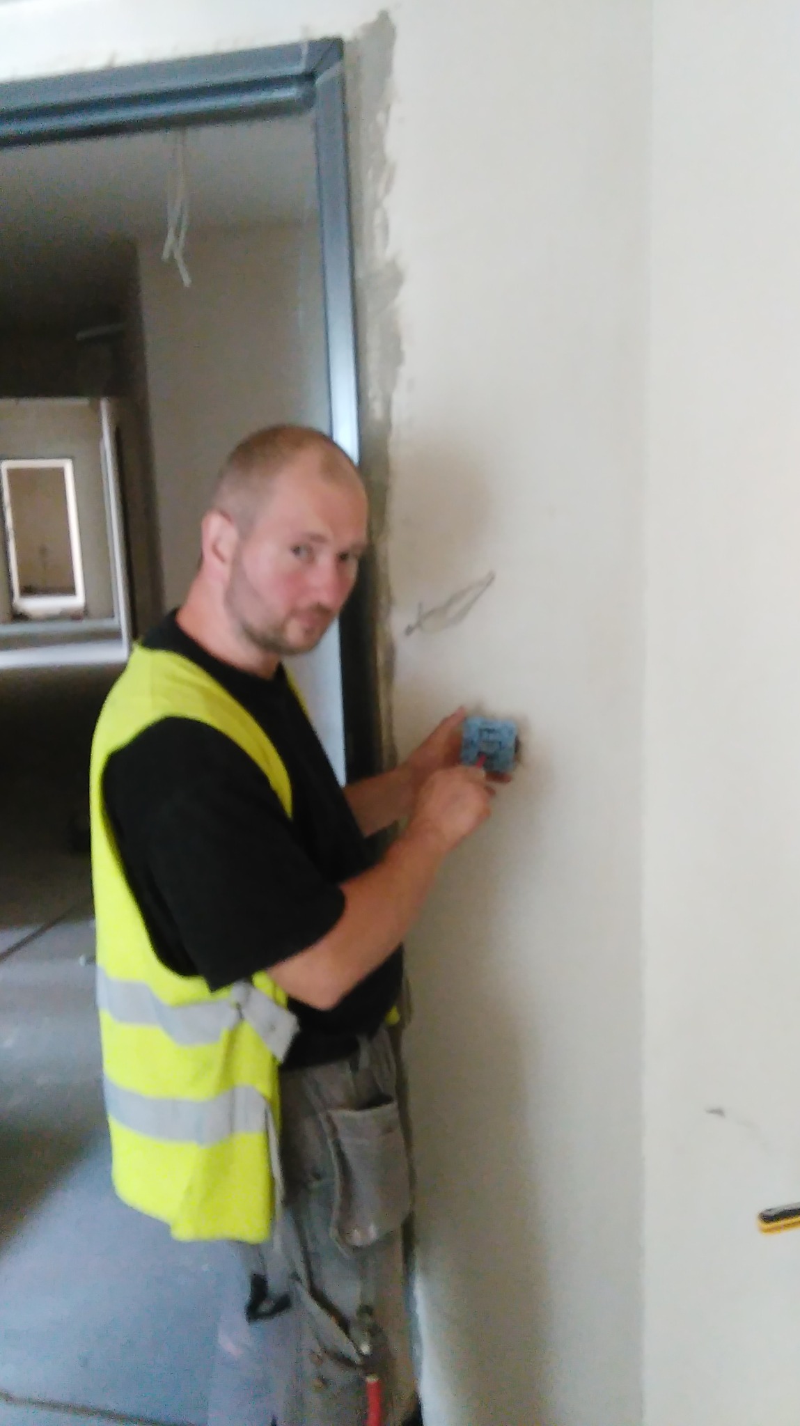 Elektryk w kamizelce odblaskowej montuje gniazdko elektryczne w ścianie podczas remontu mieszkania, widoczne przewody i surowy stan wykończenia.