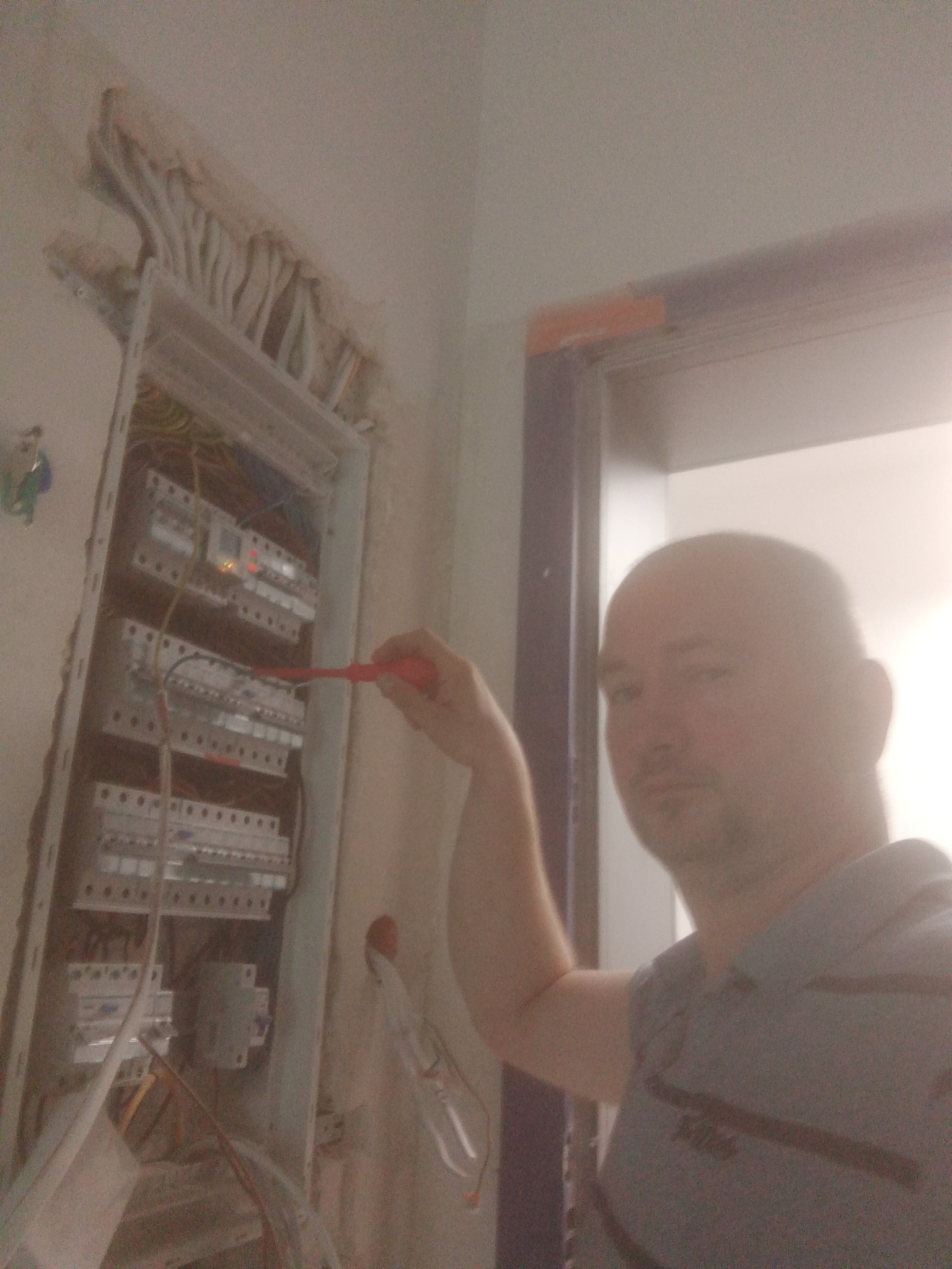 Elektryk z czerwonym śrubokrętem przy otwartej skrzynce bezpiecznikowej w trakcie prac, widoczne wystające kable ze ściany.