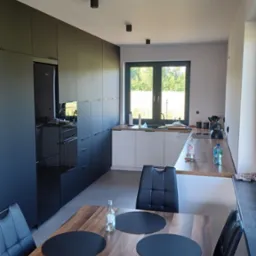 Nowoczesna kuchnia z czarnymi szafkami i sprzętem AGD, jasnymi blatami i drewnianym stołem z czarnymi podkładkami, widziana z perspektywy stołu.