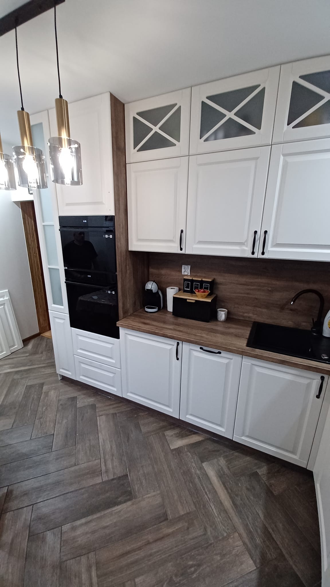 Nowoczesna kuchnia z białymi szafkami z frezowanymi frontami, drewnianym blatem i czarnymi akcentami: piekarnik, zlew i armatura. Na blacie ekspres do kawy i pojemnik na pieczywo. Podłoga wyłożona...