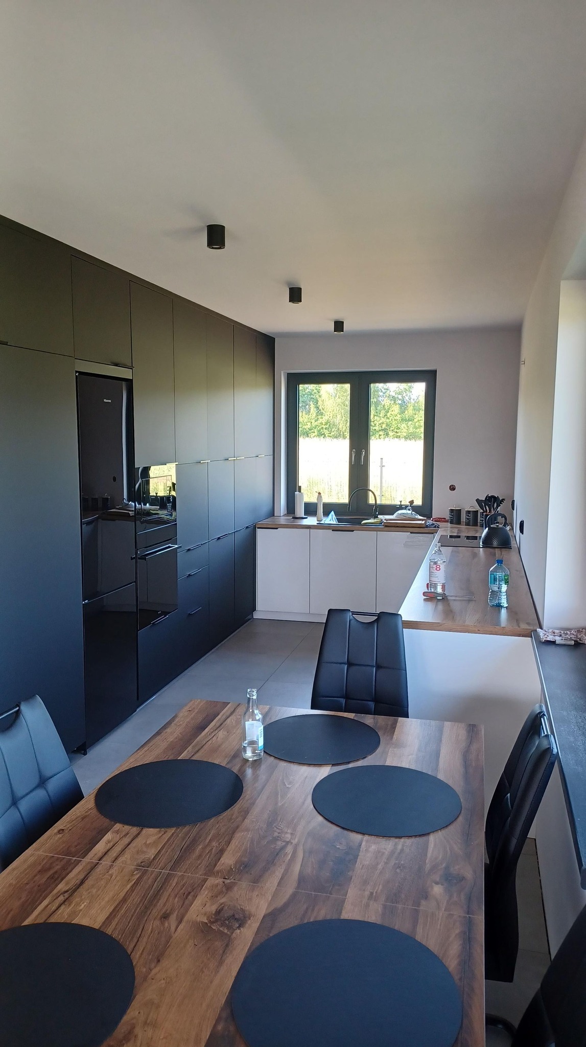 Nowoczesna kuchnia z czarnymi szafkami i sprzętem AGD, jasnymi blatami i drewnianym stołem z czarnymi podkładkami, widziana z perspektywy stołu.