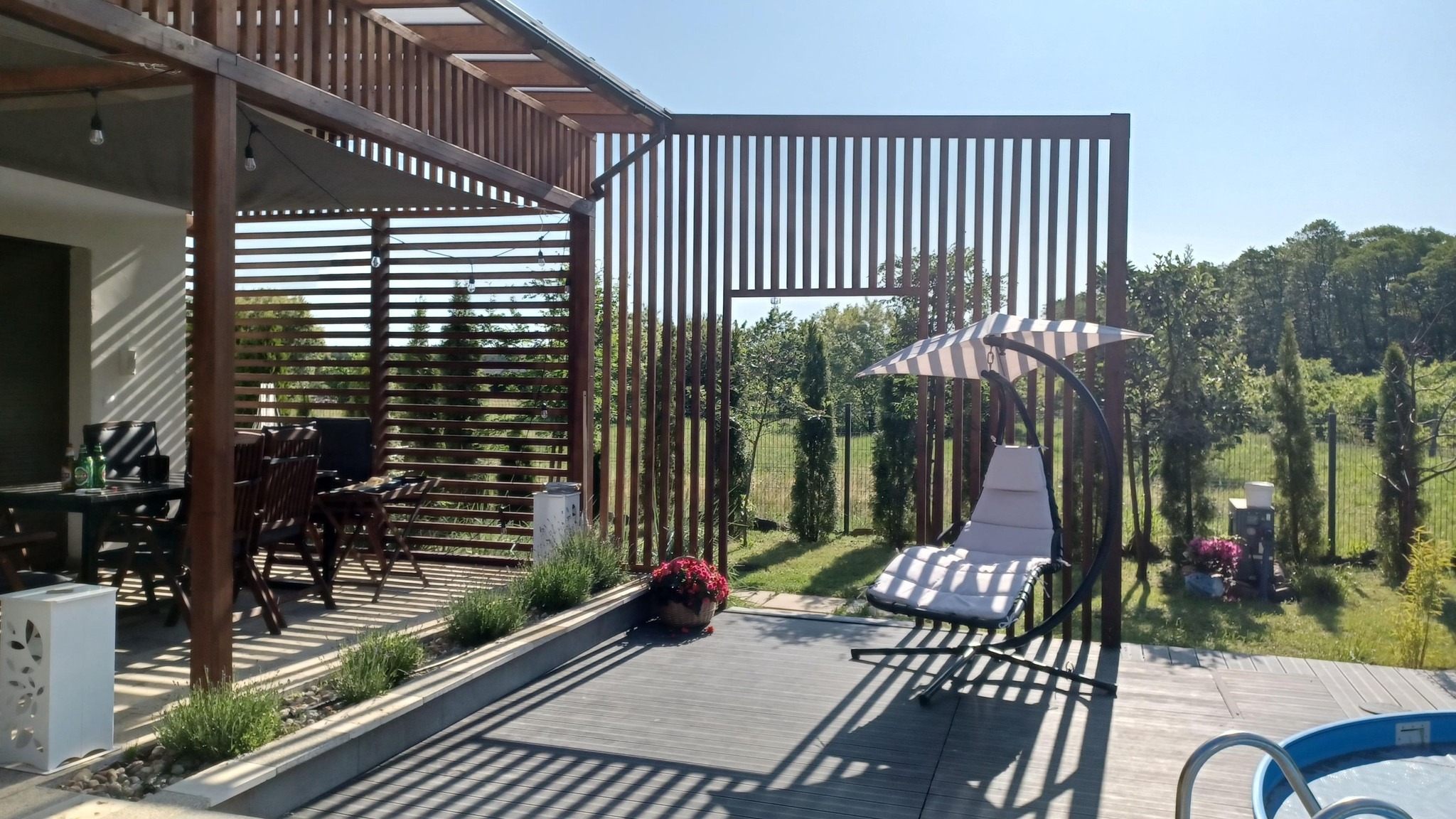 Zadaszony taras z drewnianą pergolą, szarą podłogą kompozytową i wiszącym fotelem-huśtawką, widok na ogród przez drewniane panele ażurowe.