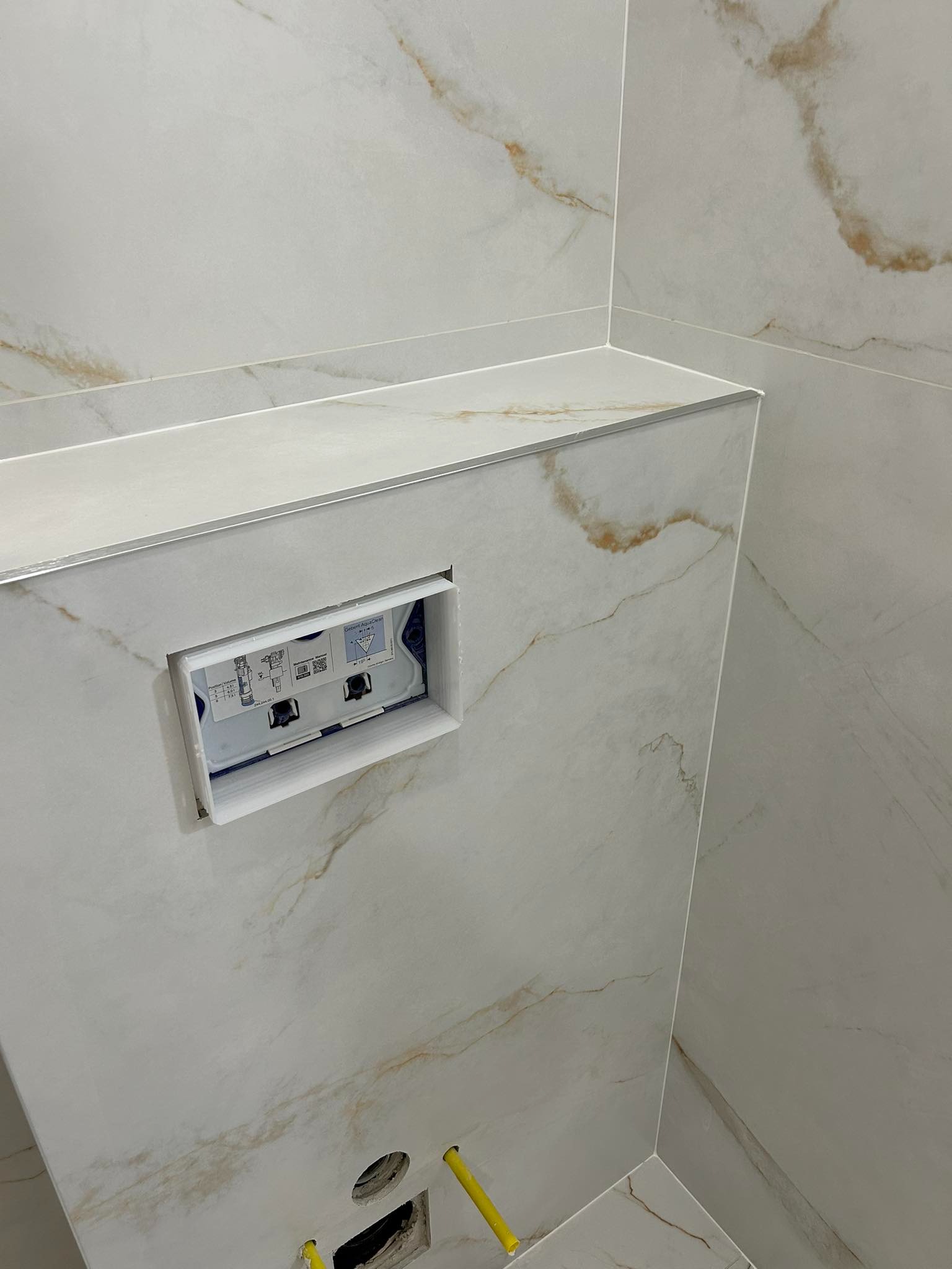 Beżowe płytki imitujące marmur na ścianie łazienki z zamontowanym stelażem podtynkowym Geberit AquaClean i wystającymi żółtymi rurkami instalacji wodnej.