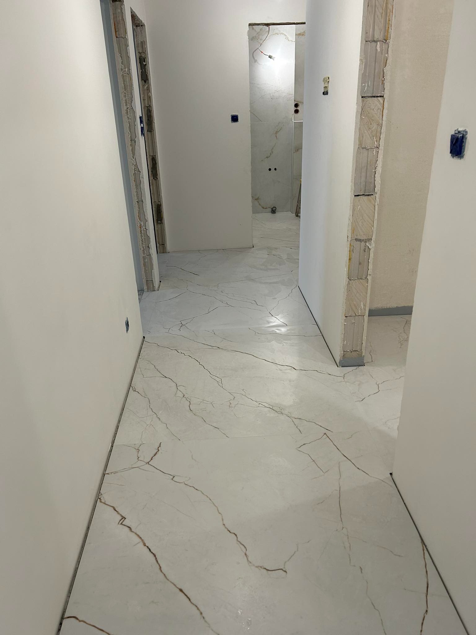Długi korytarz w trakcie remontu z nową, białą podłogą z efektem marmuru, widoczne niedokończone ościeżnice drzwiowe i instalacje elektryczne.