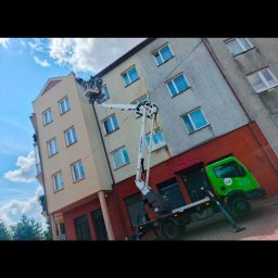 Bartosz Prelewski DomyjDach - Pracownicy w koszu podnośnika na tle bloku. Wykonywane są prace na wysokościach przy elewacji budynku. Widoczny zielony samochód-podnośnik z wysięgnikiem.