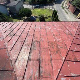 Bartosz Prelewski DomyjDach - Czerwony dach z blachy z widocznymi śladami użytkowania, z perspektywy góry, z fragmentem butów w rogu. W tle zabudowa mieszkalna i zieleń.