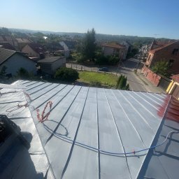 Bartosz Prelewski DomyjDach - Szary, metalowy dach z widoczną częścią pomalowaną na czerwono. W tle zabudowa miejska i błękitne niebo. Widoczne zabezpieczenia na dachu.