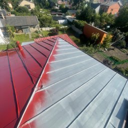 Bartosz Prelewski DomyjDach - Częściowo pomalowany na czerwono blaszany dach domu jednorodzinnego, z widoczną szarą farbą podkładową i narzędziami malarskimi. W tle zabudowa mieszkalna.