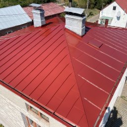 Bartosz Prelewski DomyjDach - Czerwony, metalowy dach po renowacji z dwoma kominami, widok z góry. Nowe orynnowanie w kolorze dachu. W tle budynki mieszkalne i zieleń.