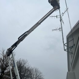 Bartosz Prelewski DomyjDach - Pracownik w podnośniku koszowym naprawia antenę na budynku. Widok z dołu na pochmurne niebo, drzewa i fragment elewacji. Praca na wysokości.