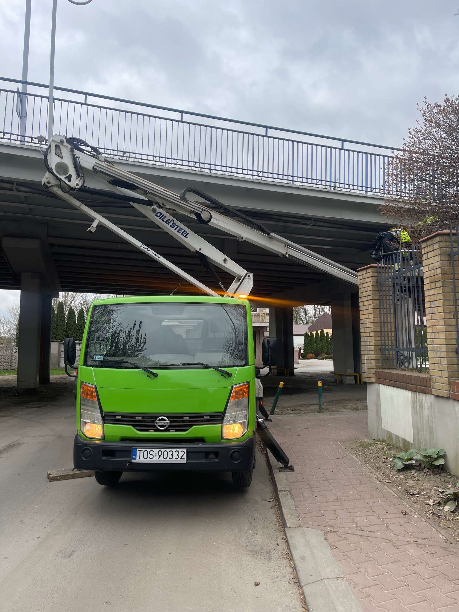 Zielony podnośnik koszowy OIL&STEEL z wysięgnikiem pod mostem. Pracownik w żółtej kamizelce na górze. Tablica rejestracyjna TOS 90332.