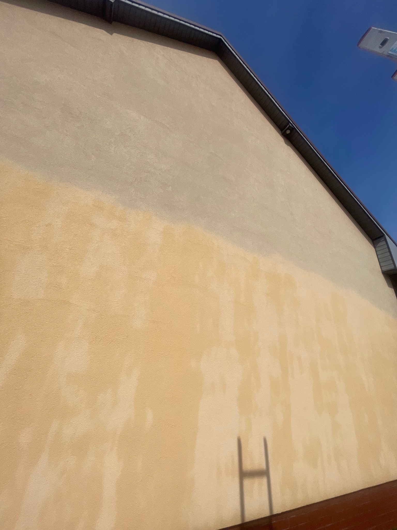 Częściowo umyta, beżowa elewacja budynku z widocznym cieniem drabiny. Kontrast czystej i zabrudzonej powierzchni. Górna część elewacji jest jaśniejsza.