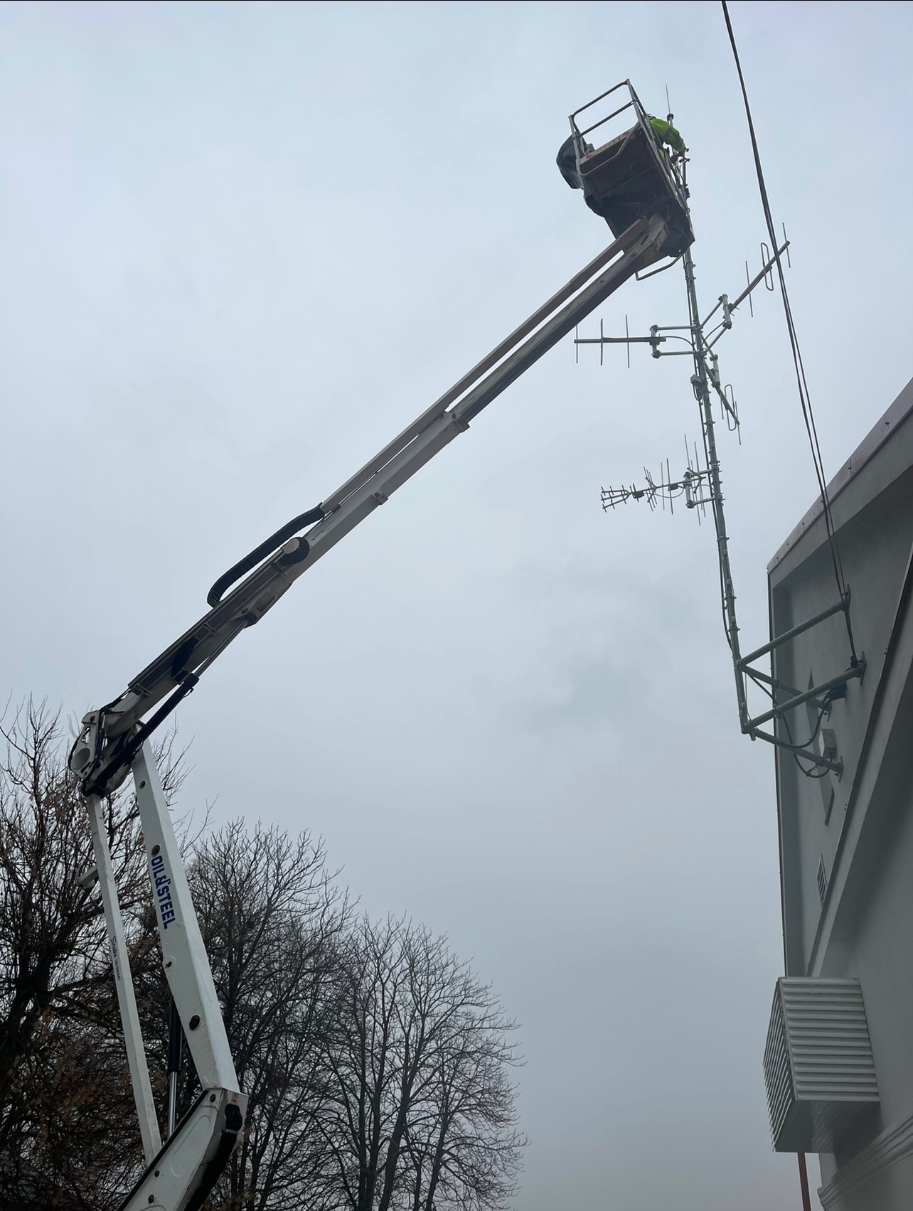 Pracownik w podnośniku koszowym naprawia antenę na budynku. Widok z dołu na pochmurne niebo, drzewa i fragment elewacji. Praca na wysokości.