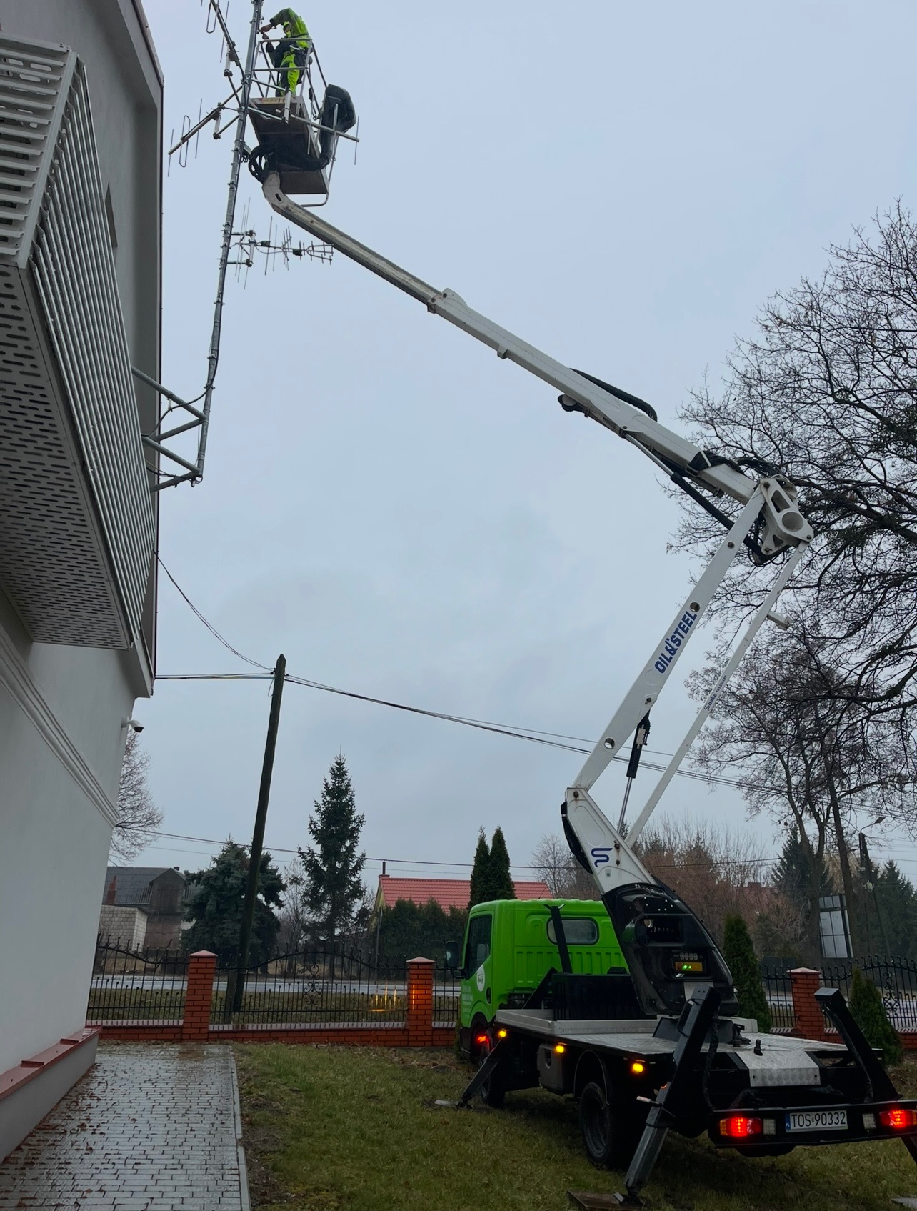 Pracownik w podnośniku koszowym przytwierdza antenę do elewacji budynku. Widoczny zielony samochód z podnośnikiem na tle zabudowy jednorodzinnej.