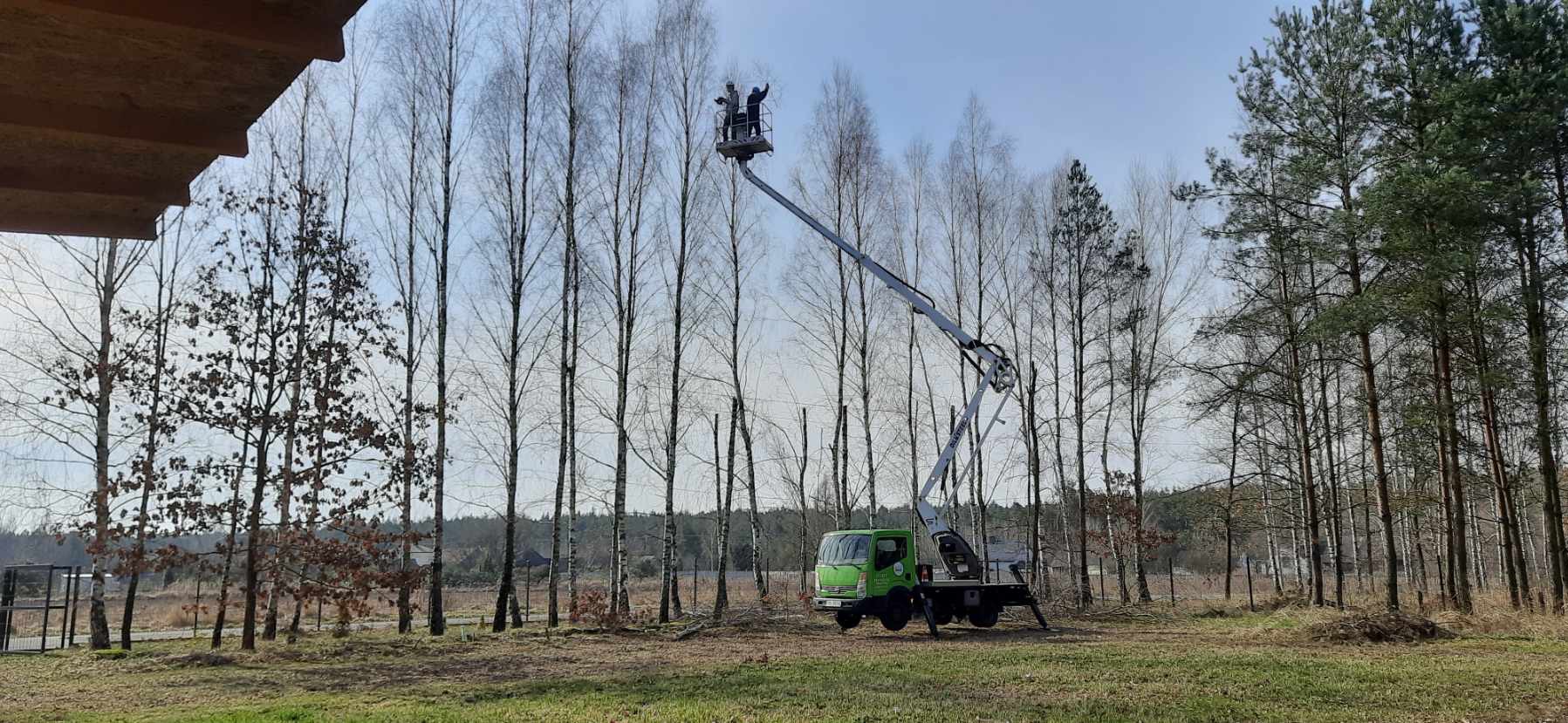 Zielony podnośnik koszowy na tle drzew, dwóch pracowników w koszu podnosi ręce. Widoczne ścięte gałęzie. Praca w terenie otwartym, w tle las i błękitne niebo.