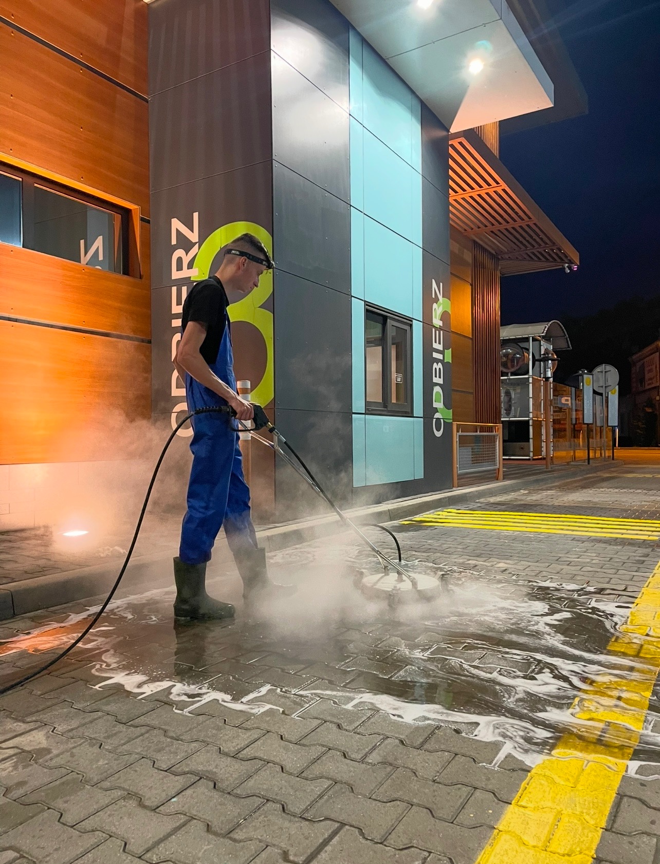 Pracownik w kombinezonie czyści kostkę brukową przed budynkiem z napisem 'ODBIERZ', używając myjki ciśnieniowej z obrotową szczotką. Widoczne żółte pasy i woda.