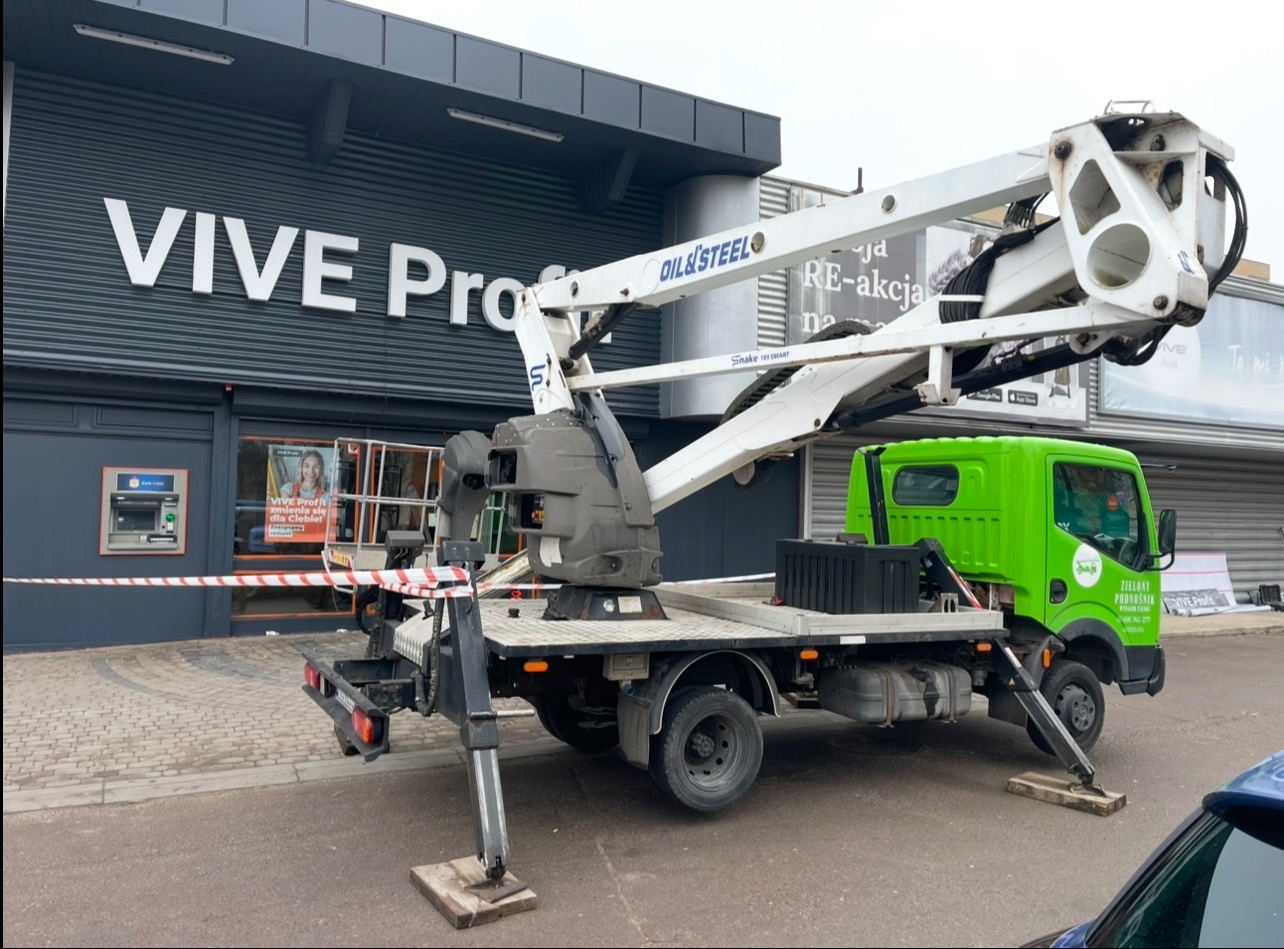 Podnośnik koszowy z wysięgnikiem, ustawiony przed budynkiem z napisem VIVE Pro. Widoczne stabilizatory i zielona kabina pojazdu. Na budynku reklama i bankomat.