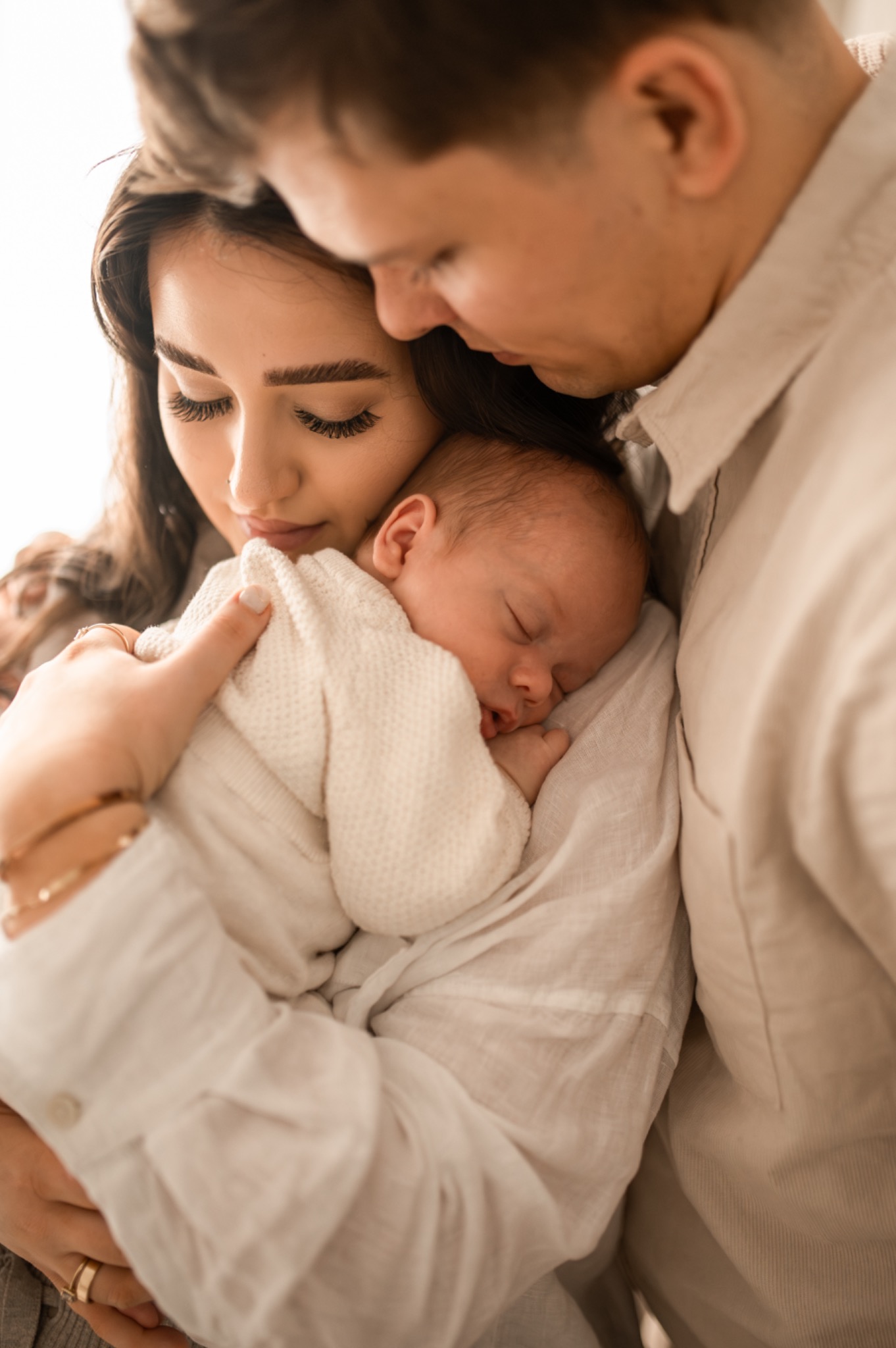 Rodzice tulący śpiącego noworodka w jasnych, naturalnych barwach, delikatne oświetlenie, widoczny pierścionek na palcu matki.