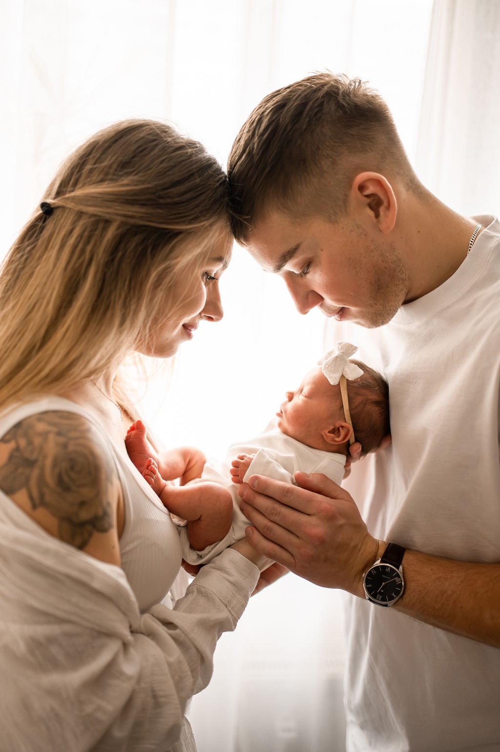 Rodzice z czułością patrzą na śpiące nowo narodzone dziecko w białym ubranku i z kokardką na głowie, mama ma tatuaż na ramieniu, tata zegarek na ręce.