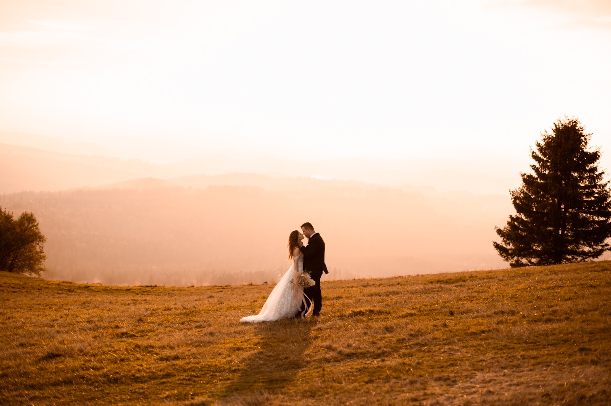 Para młoda w objęciach na wzgórzu o zachodzie słońca, w tle zarys gór i samotne drzewo iglaste.