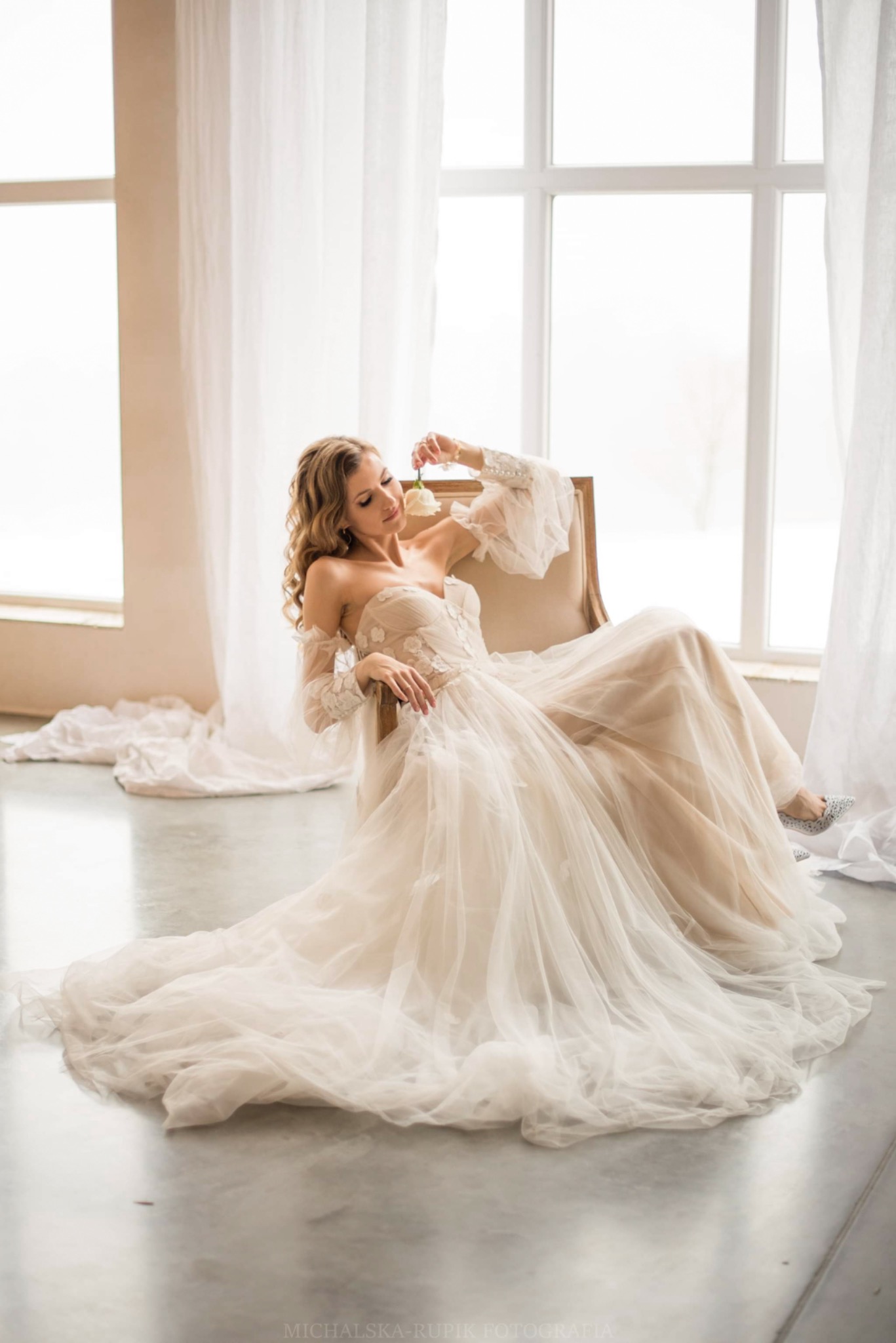 Kobieta w zwiewnej sukni ślubnej z kwiatem, siedząca w fotelu na tle okna z białymi zasłonami.