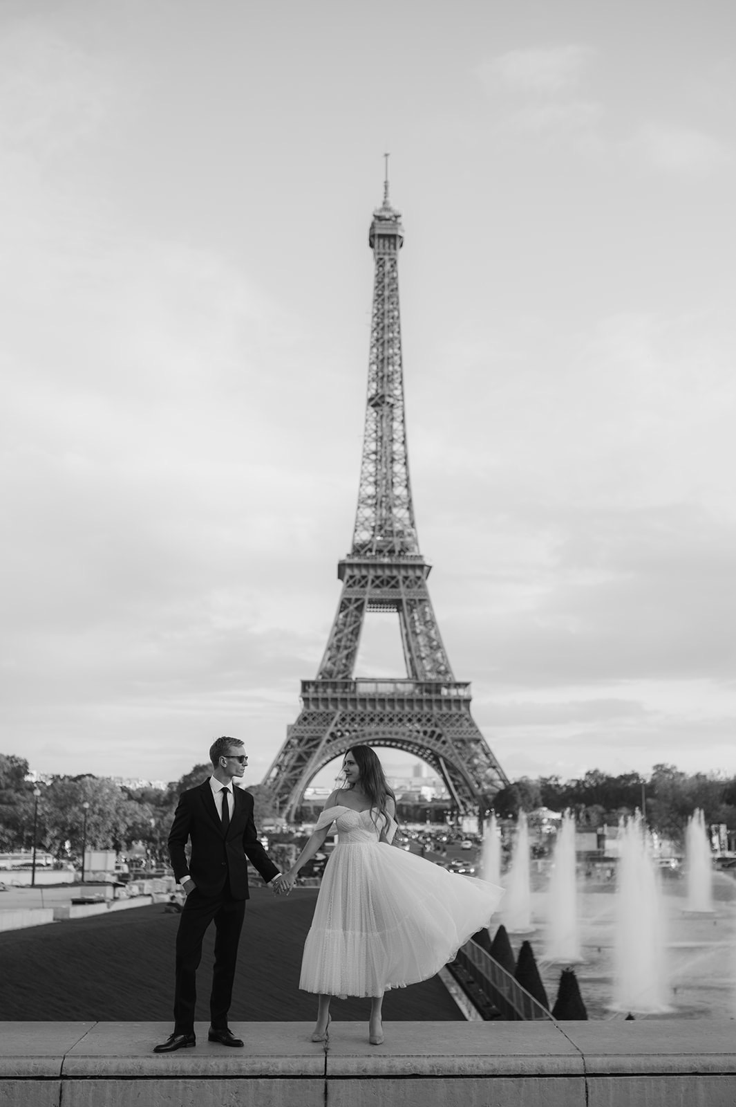 Czarno-białe zdjęcie pary trzymającej się za ręce na tle wieży Eiffla i fontann, mężczyzna w garniturze i okularach przeciwsłonecznych, kobieta w białej sukience.