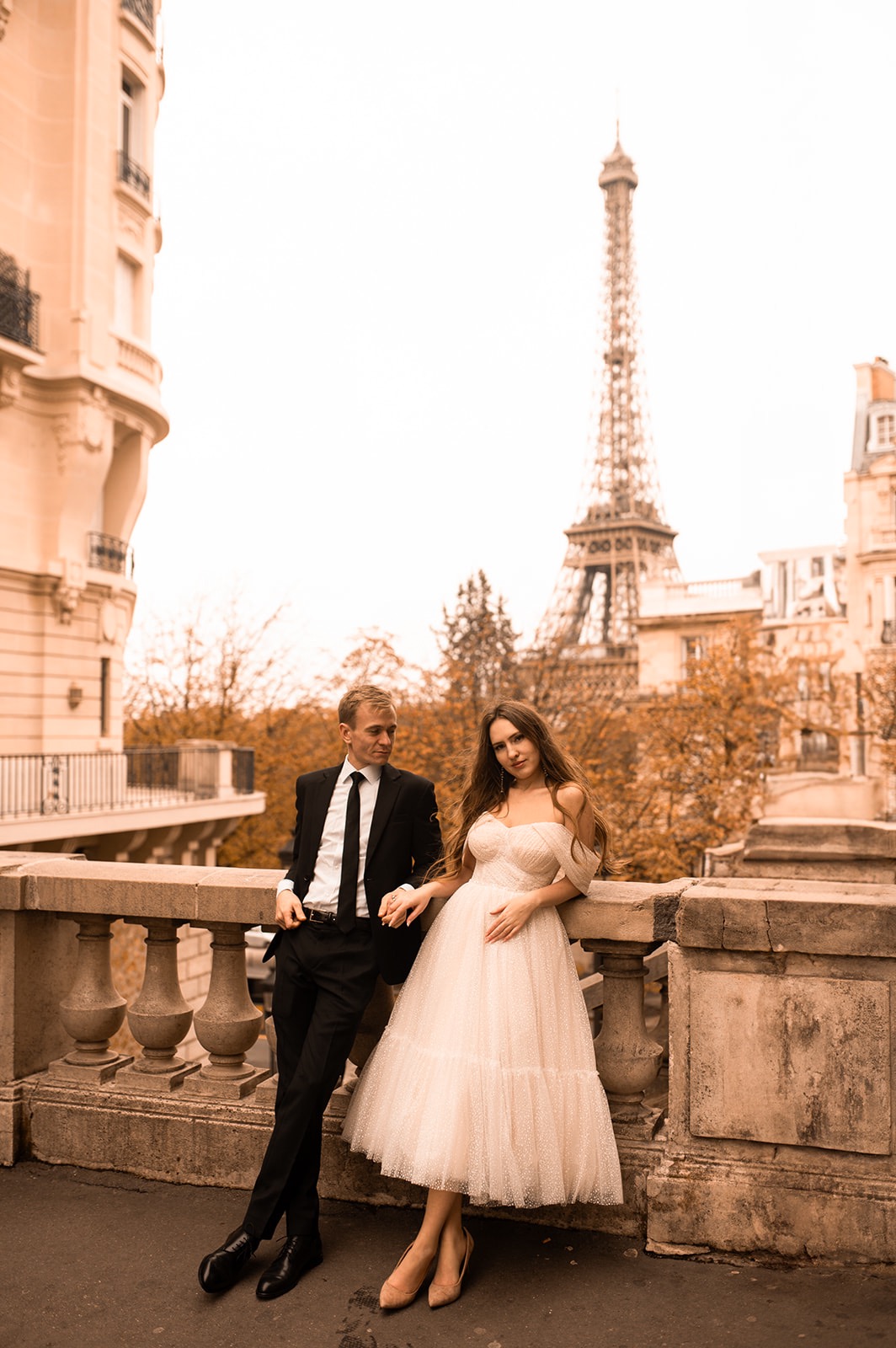 Elegancka para w Paryżu, mężczyzna w czarnym garniturze, kobieta w tiulowej sukience, w tle wieża Eiffla i kamienice.