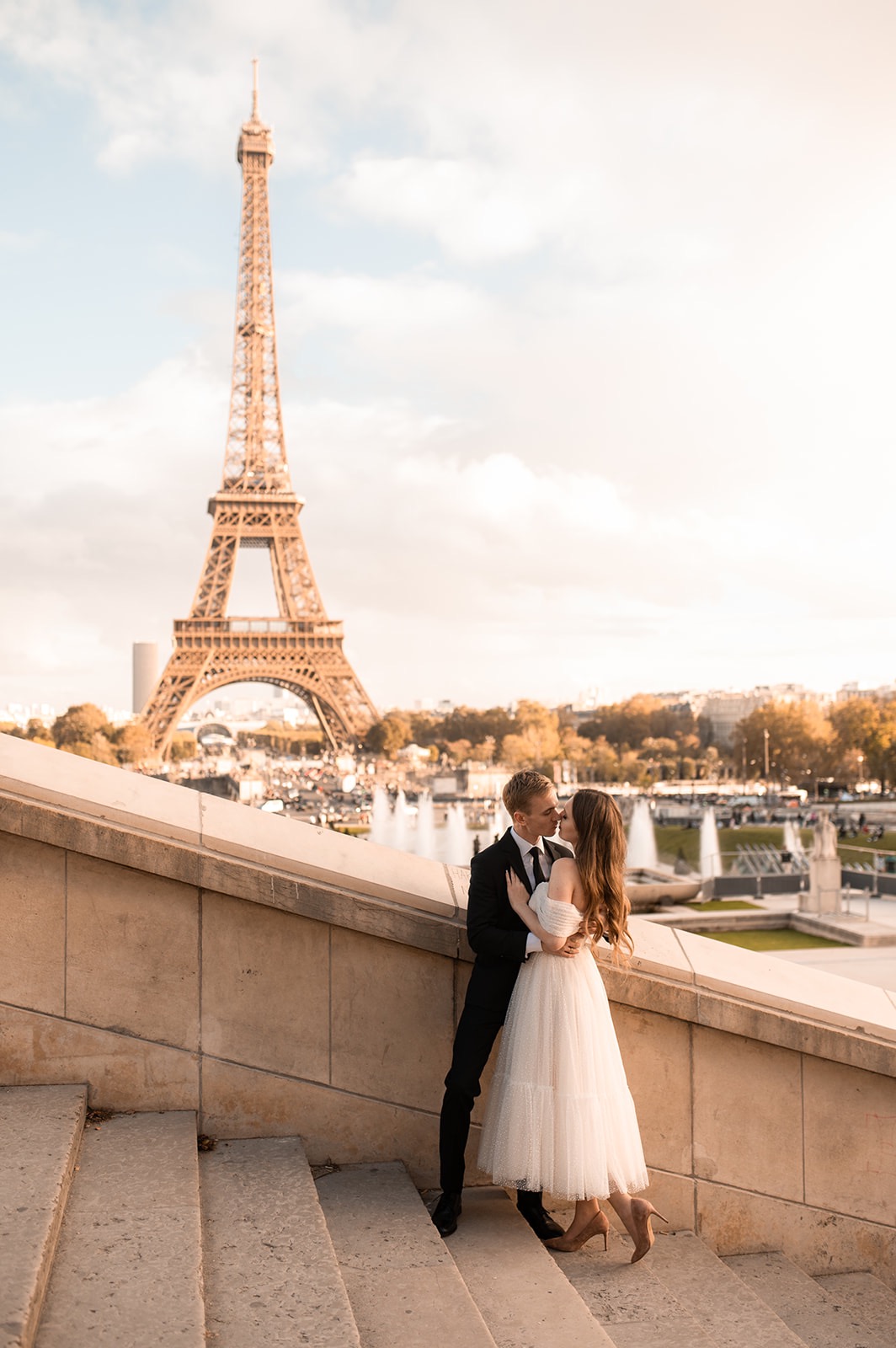 Zakochana para całuje się na tle wieży Eiffla w Paryżu, ona w zwiewnej sukience midi z tiulu, on w czarnym garniturze, ujęcie z dołu schodów.