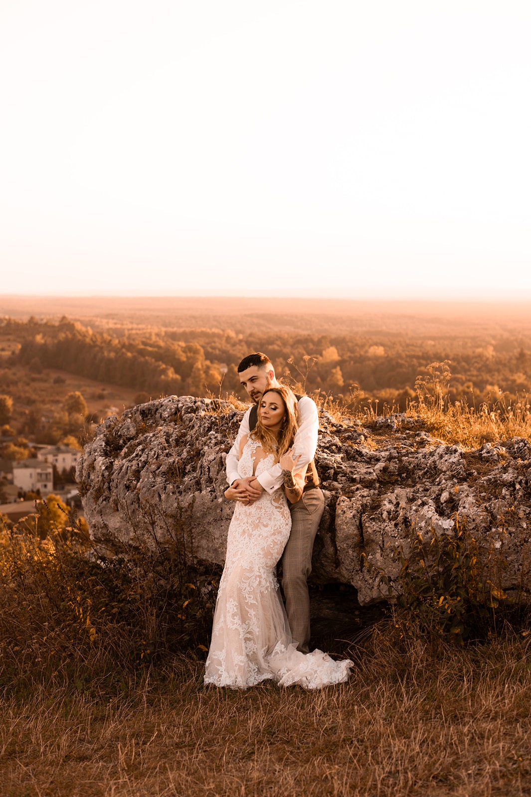 Para narzeczeńska w Orzeszu pozuje na tle malowniczego krajobrazu w złotej godzinie, ona w koronkowej sukni ślubnej z długim rękawem, on w kamizelce i spodniach w kratę, opierają się o duży, szary...