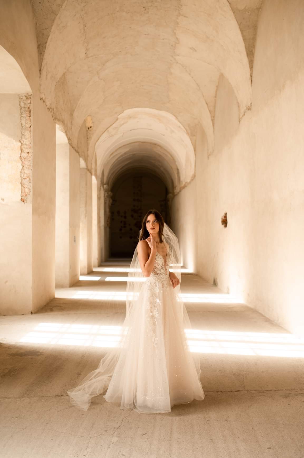 Panna młoda w koronkowej sukni ślubnej i welonie pozuje w długim, jasnym korytarzu z łukowym stropem, oświetlonym promieniami słonecznymi wpadającymi przez boczne okna.