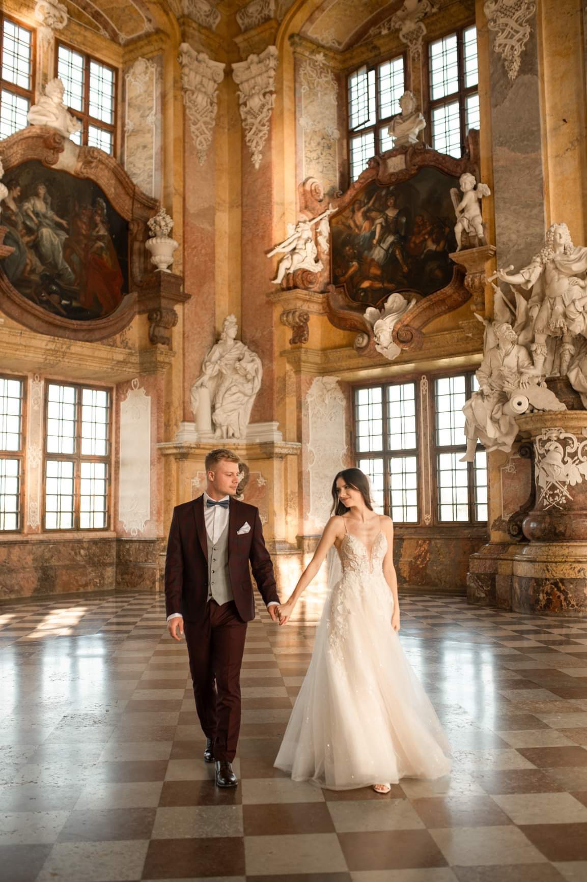Para młoda trzymająca się za ręce w barokowym wnętrzu z marmurową podłogą w szachownicę, rzeźbami i obrazami na ścianach.