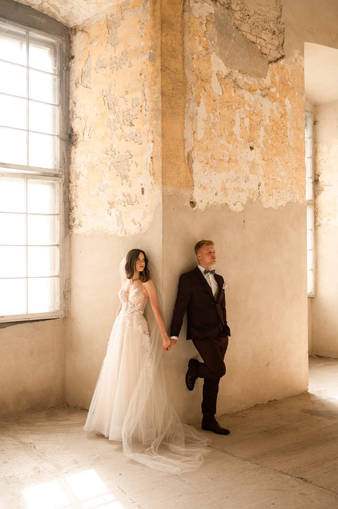 Para młoda trzymająca się za ręce, oparta o narożnik ściany z łuszczącą się farbą w opuszczonym budynku z oknami.