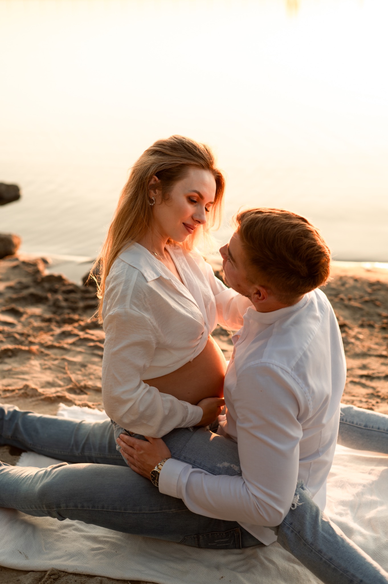 Ciężarna kobieta w białej koszuli siedzi na piasku w objęciach mężczyzny, oboje patrzą na siebie z czułością, w tle zachód słońca nad wodą.