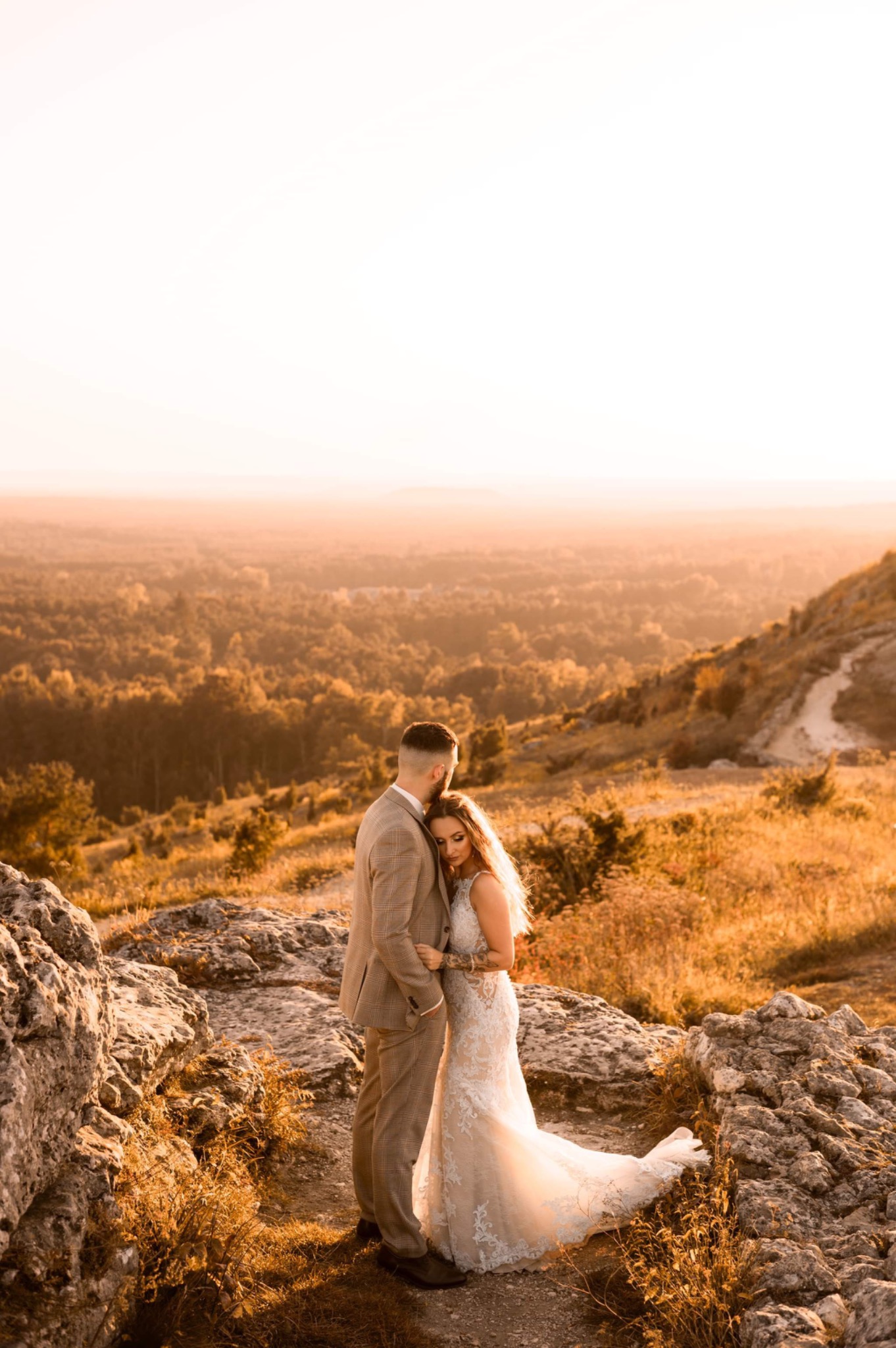Para w strojach ślubnych obejmuje się na skalistym wzgórzu o zachodzie słońca, z widokiem na zalesioną dolinę w oddali.