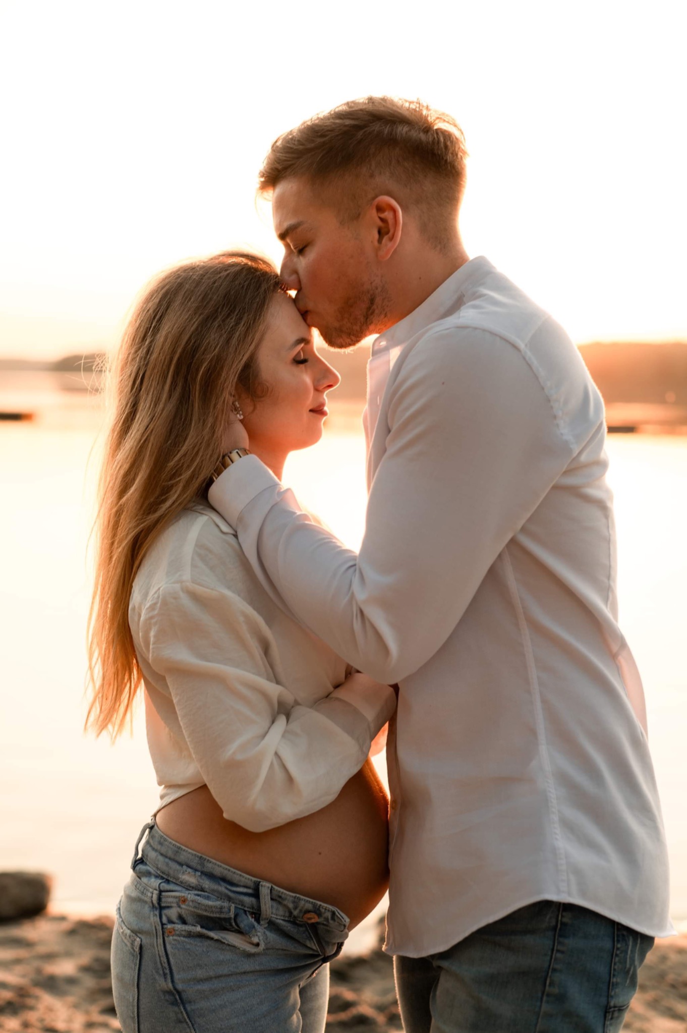 Zakochana para na plaży o zachodzie słońca; mężczyzna całuje kobietę w czoło, widoczny ciążowy brzuszek kobiety.