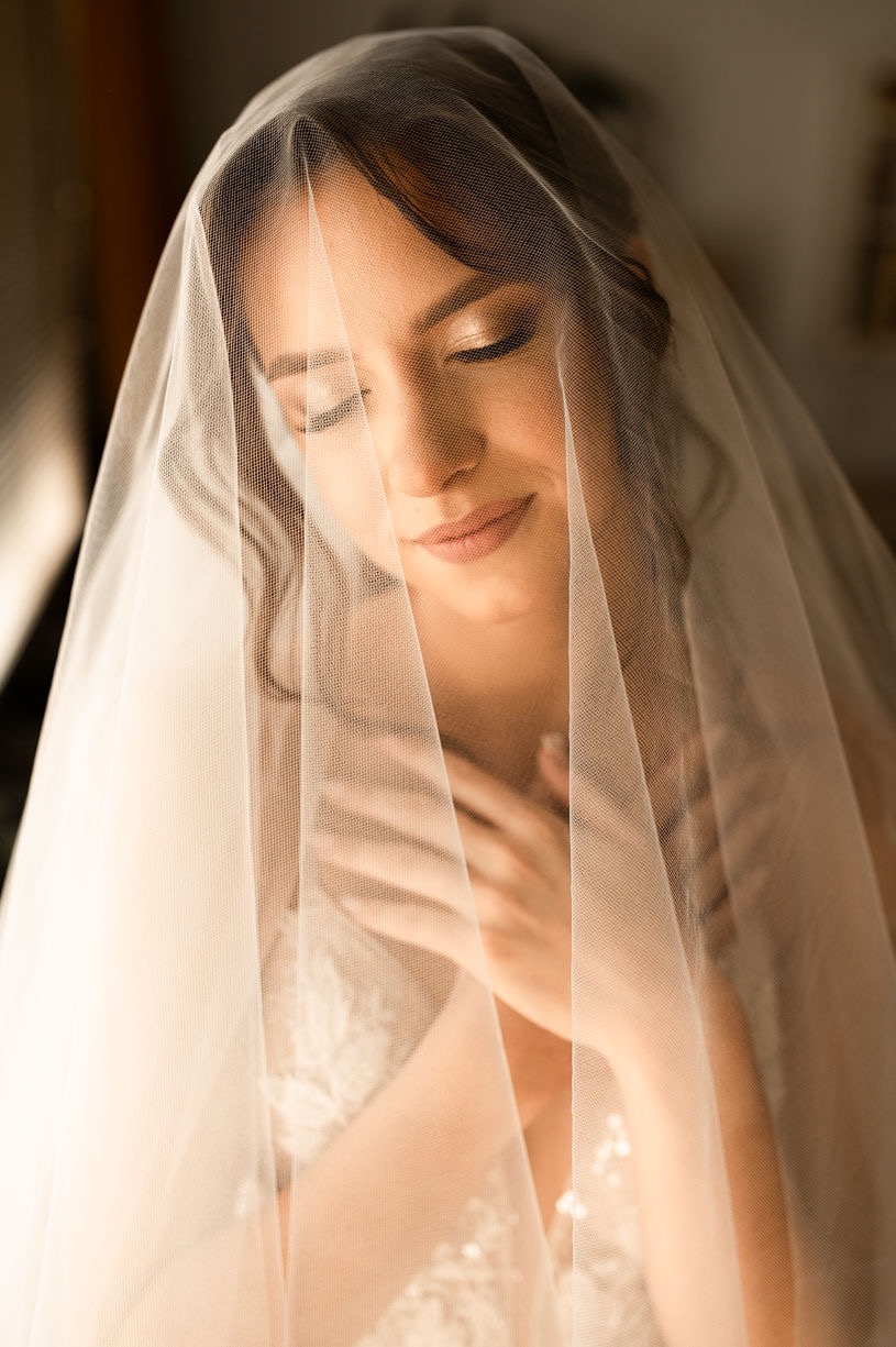 Uśmiechnięta panna młoda z zamkniętymi oczami, delikatny makijaż, częściowo zasłonięta welonem, dłonie na dekolcie sukni ślubnej.