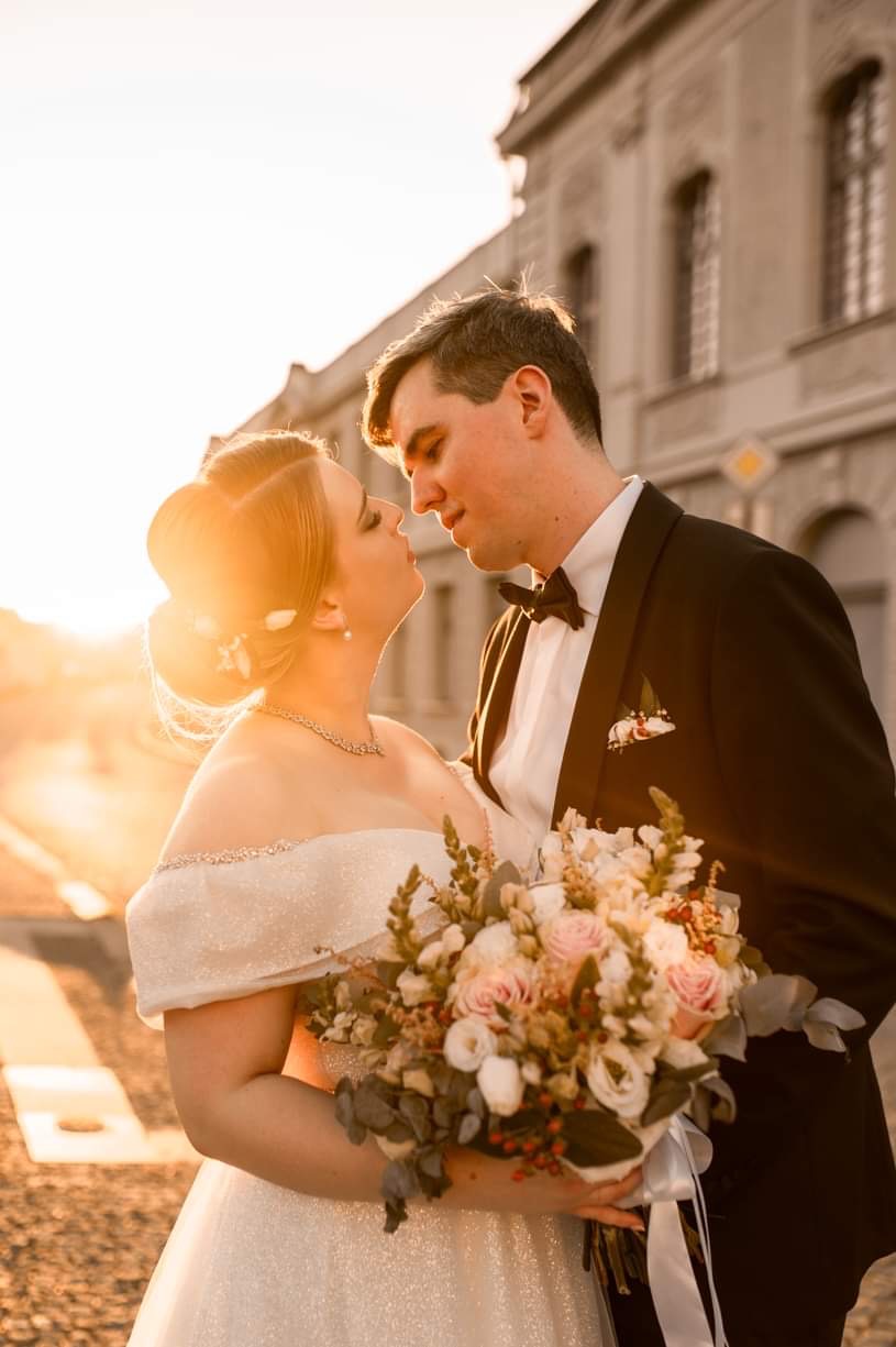Para młoda, delikatnie oświetlona zachodzącym słońcem, w eleganckich strojach ślubnych, trzyma bukiet z różowych i białych róż na tle klasycznej architektury miejskiej.